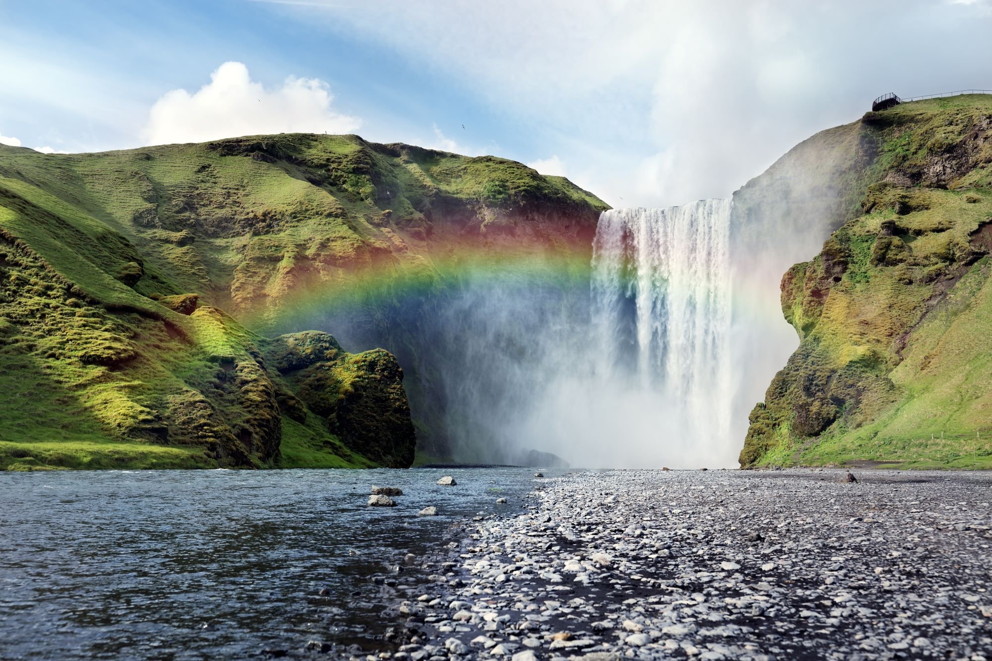 A scenic view of a large waterfall cascading down a cliff surrounded by lush green hills, with a rainbow forming in the mist near the waterfall.