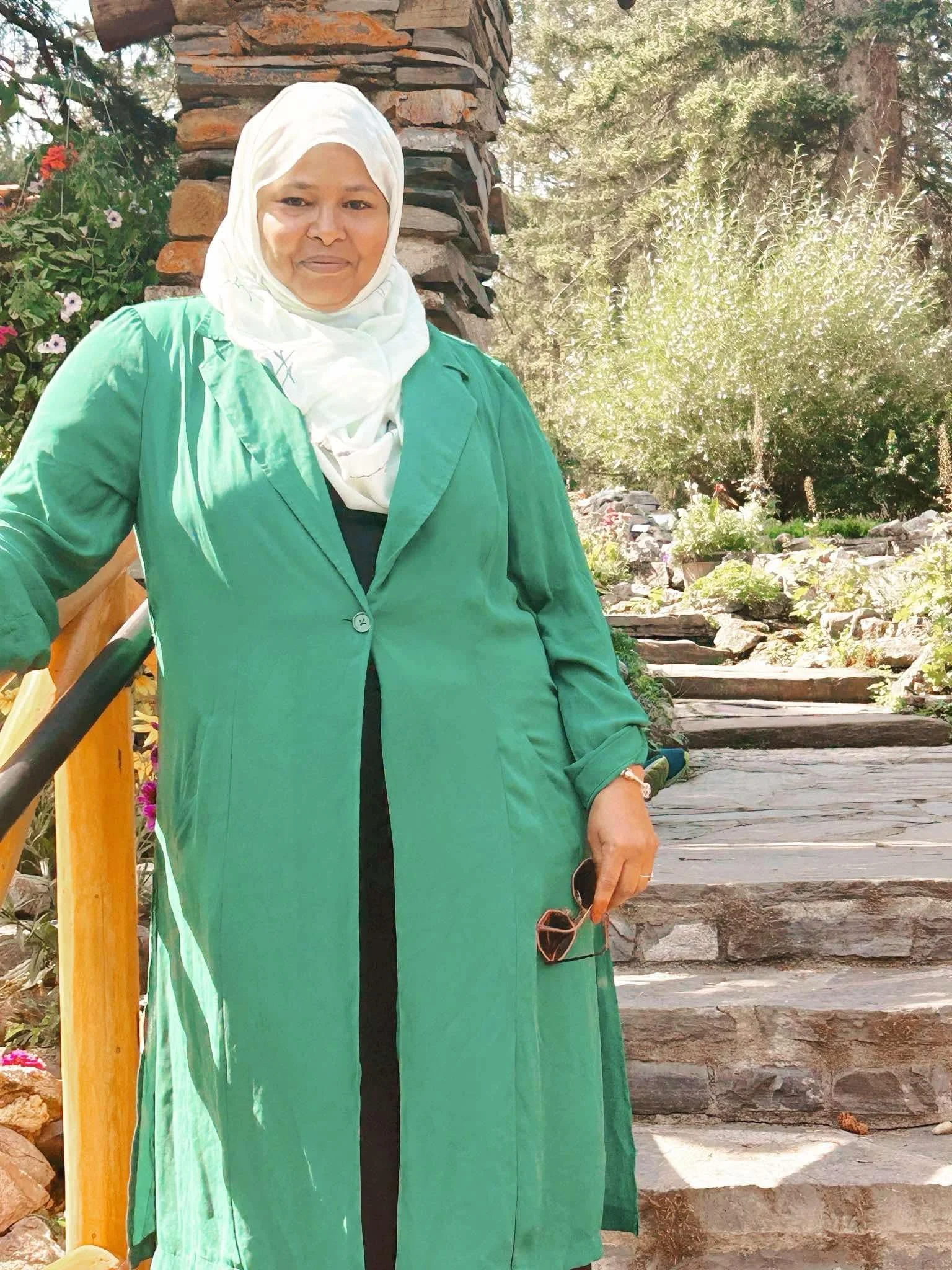 A woman wearing a white hijab and a green coat, standing outdoors on stone steps with trees and plants in the background.