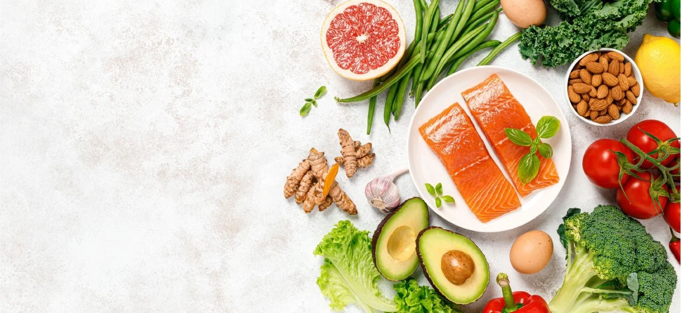 Assorted healthy foods including salmon, avocados, tomatoes, green beans, kale, broccoli, almonds, eggs, a grapefruit, and turmeric on a light background.