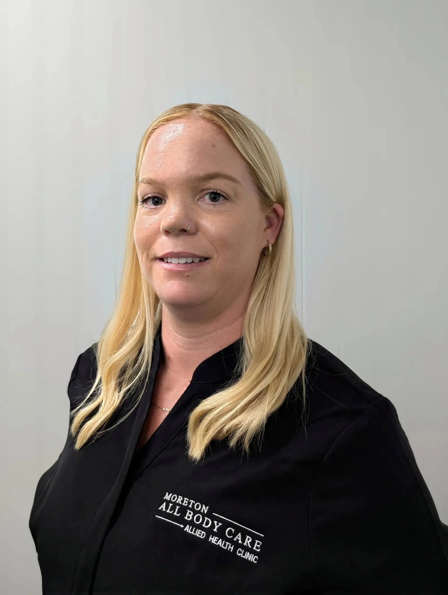 A woman with blonde hair and a nose ring, wearing a black shirt with the logo 'Moreton All Body Care Allied Health Clinic,' standing against a plain white wall.