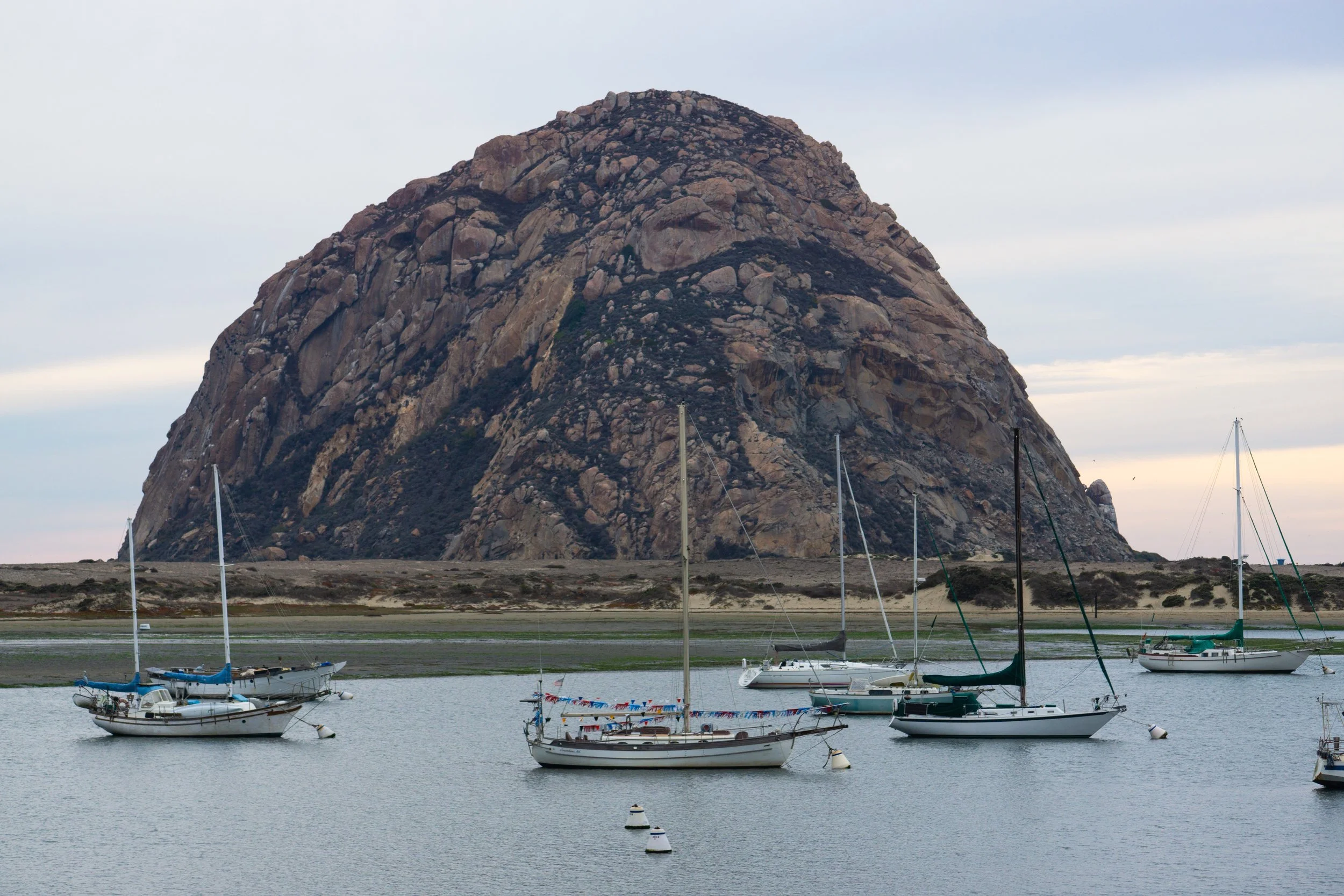 Several sailboats anchored in a calm harbor with Morro Rock in the background.