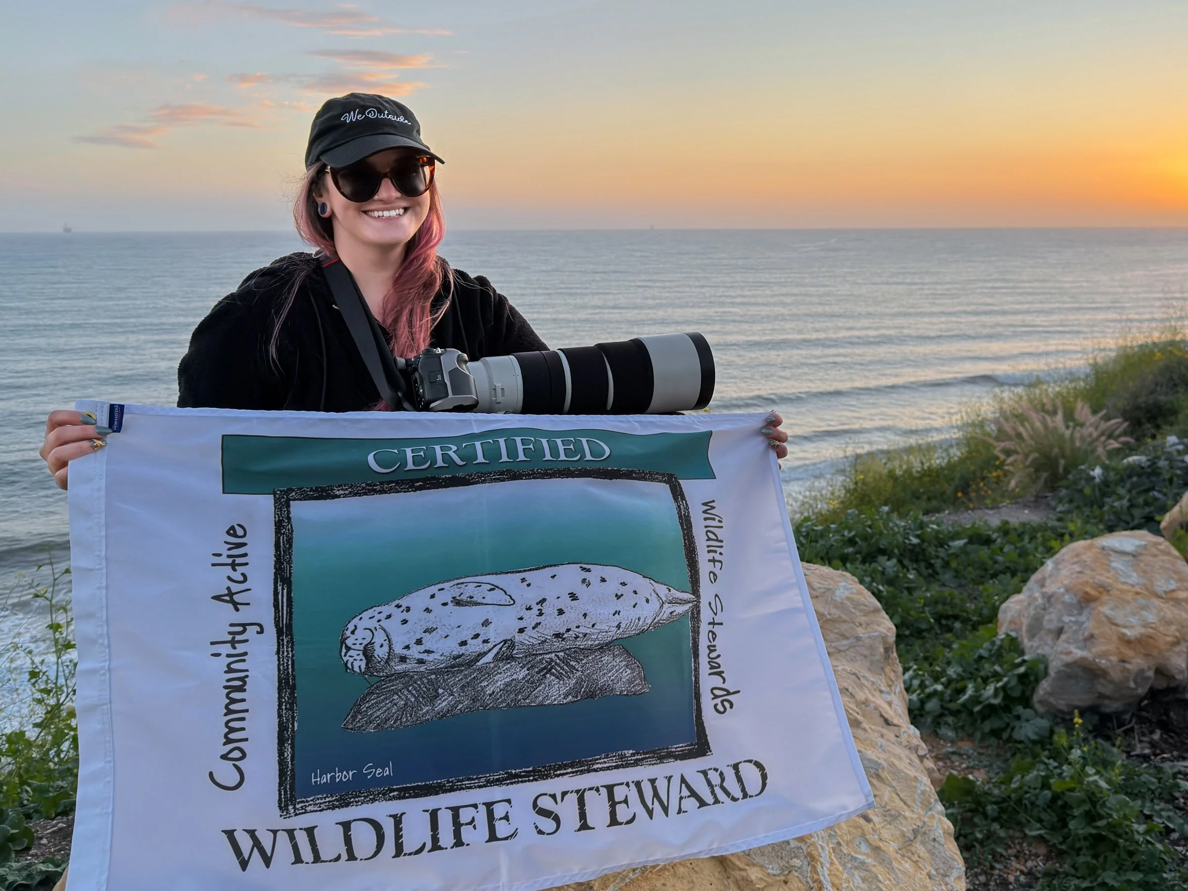 Alexis Noelle holds a Sea Otter Savvy CAWS (Certified Wildlife Steward) flag, with a sunset in the background.