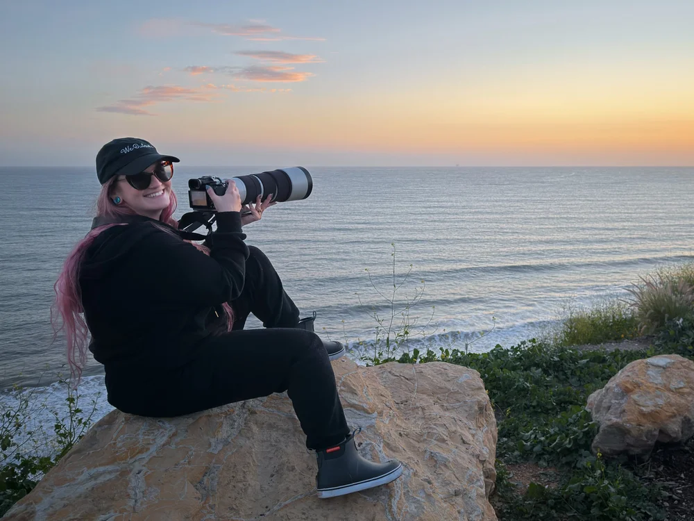 Alexis Noelle Galasso on coastal rocks at sunset with a Sony A1 II and telephoto lens.