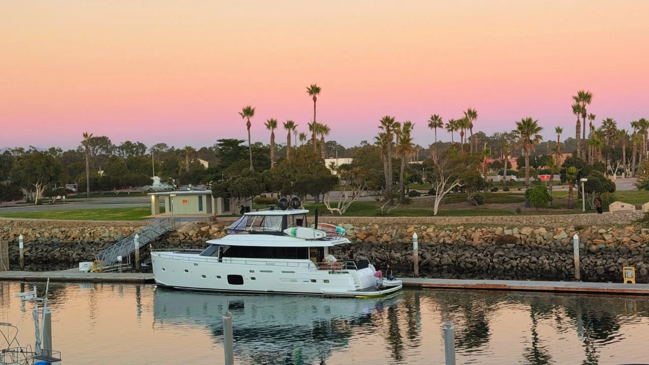 Sunset over Channel Islands Harbor from the Coastal Café livestream, looking across the channel toward the dock and palm-lined shore.