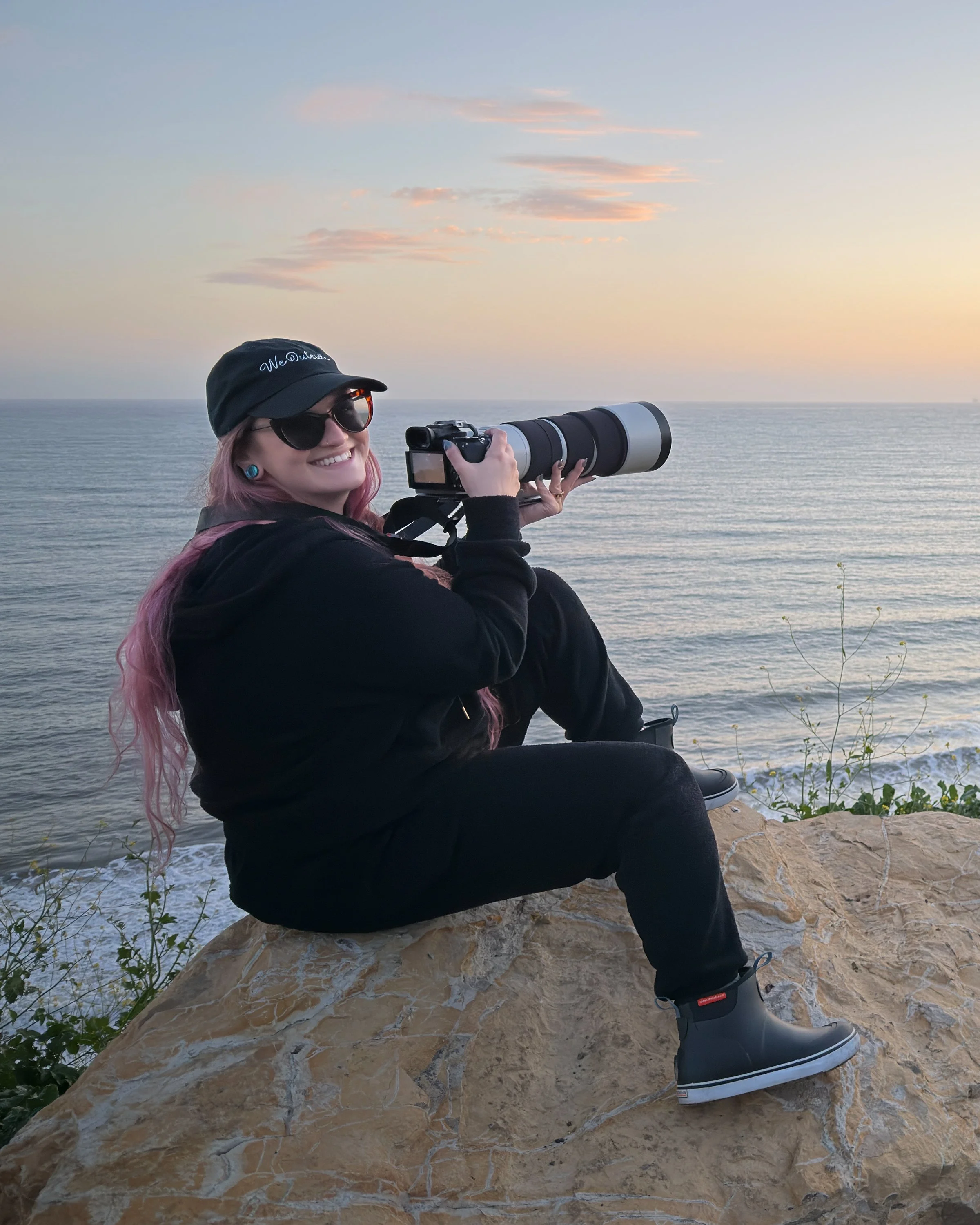 Person sitting on rocks by the ocean at sunset, taking a photograph with a large camera lens. Wearing sunglasses, a black cap, and black clothing, smiling and looking towards the water with the sunset sky in the background.