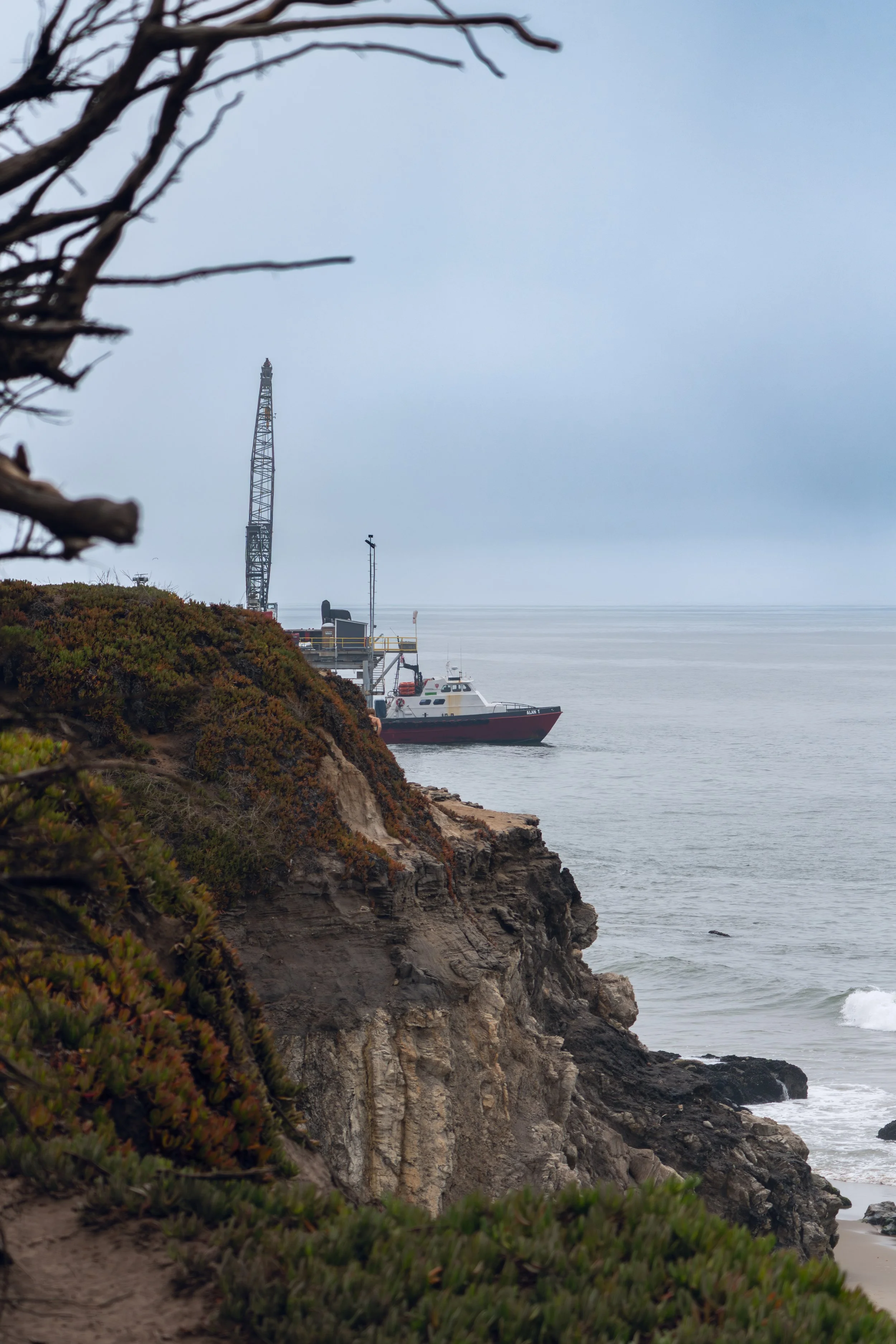 Cliffside view of a boat docked near the rocky shoreline, with overcast sky and sparse vegetation in the foreground.