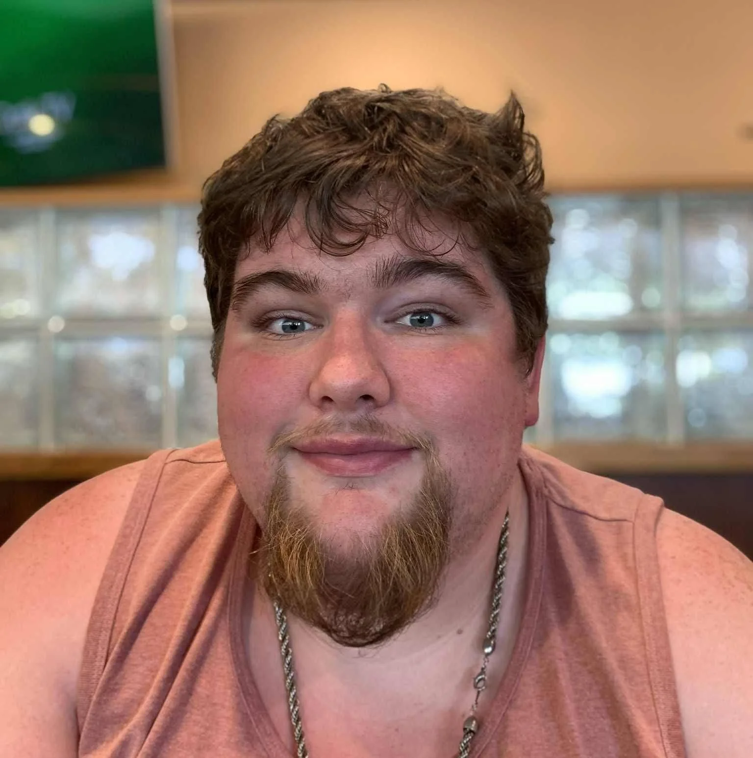 A photo of Jesse from the chest up in front of a blurred background. He has a neutral expression and has light brown hair and a brown beard and moustache. He is wearing a coral tank top and a necklace.