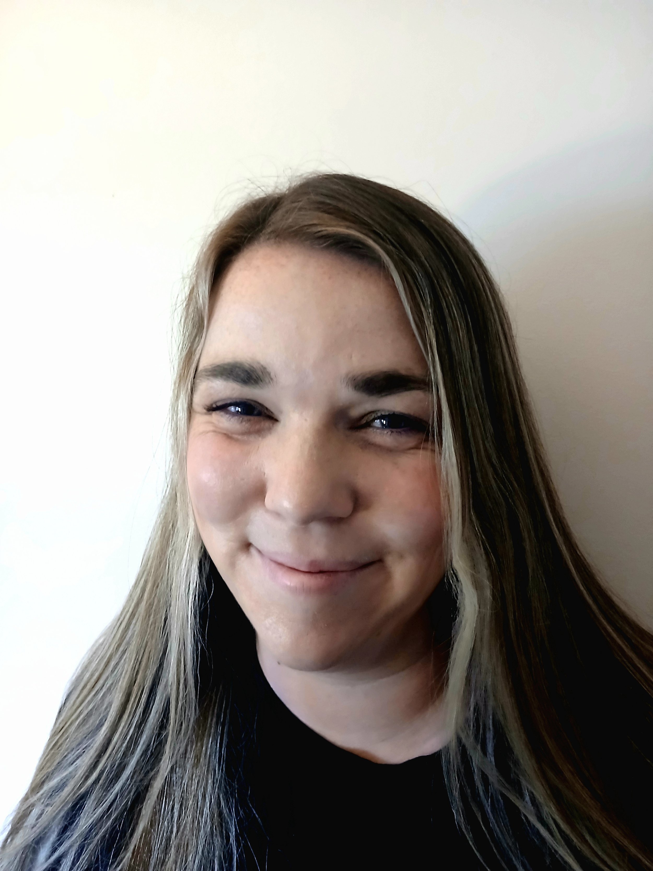 Photo of Kelly from the shoulders up with a white background. She is smiling and wearing a plain black shirt. She has long light brown/blonde hair, blue eyes and freckles.