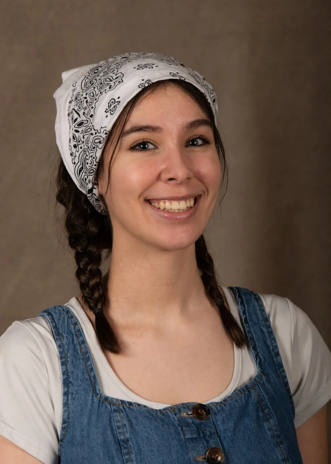 A photo of Autumn from the shoulders up in front of a brown background. She is wearing a white shirt and blue overalls and a white bandana on her head. She is smiling and has long braided brown hair and brown eyes.