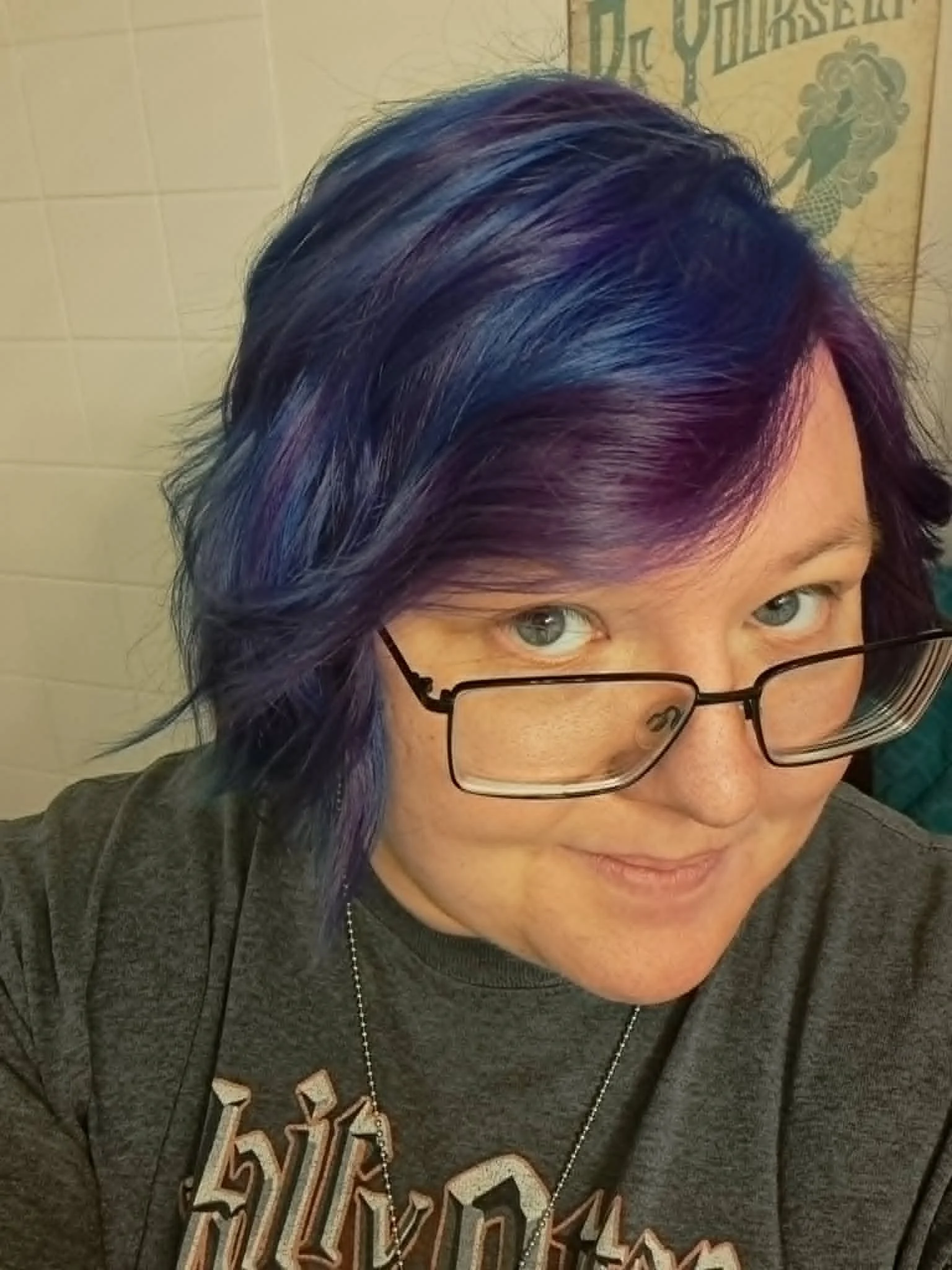 A photo of Brittany available a white brick background from the shoulders up. She is facing the camera at an angle and has dark blue and purple hair, blue eyes, and is wearing black framed glasses. She is wearing a gray t-shirt.