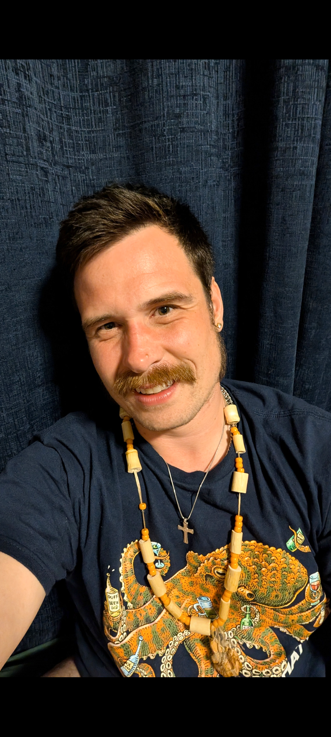 A photo of Daniel from the shoulders up in front of a navy blue background. He is smiling and wearing a navy blue shirt and a necklace. He has short brown hair and a brown moustache.