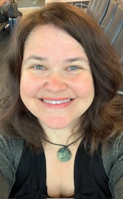 Photo of Lisa from the shoulders up with no background. She is smiling, and has medium length brown hair and blue eyes. She is weather a grey top and a necklace.