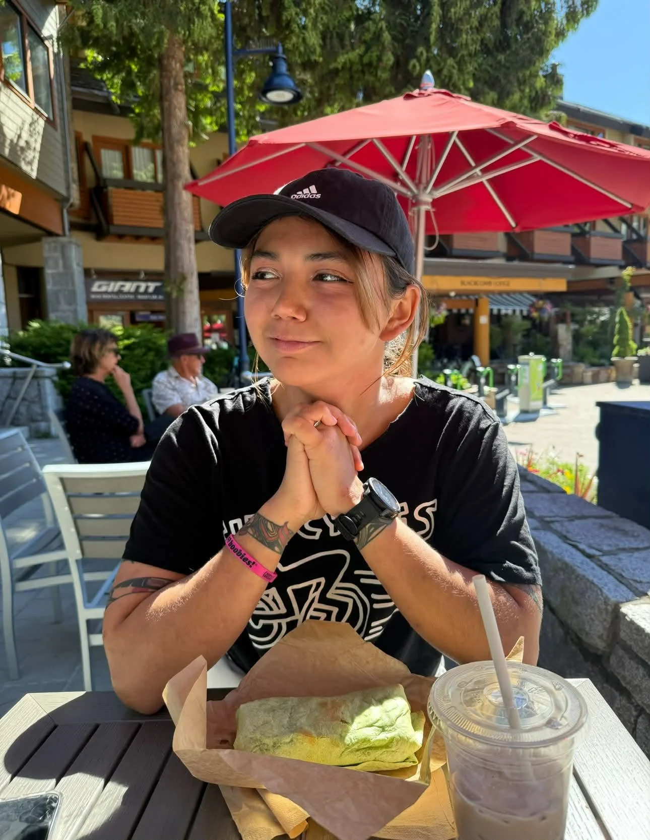 A photo of Talela from the waist up with her elbows on a table, hands clasped in front of her chest, and she is looking off to the side. She has a black baseball cap on and brown hair. There is a drink cup and a wrap in front of her.