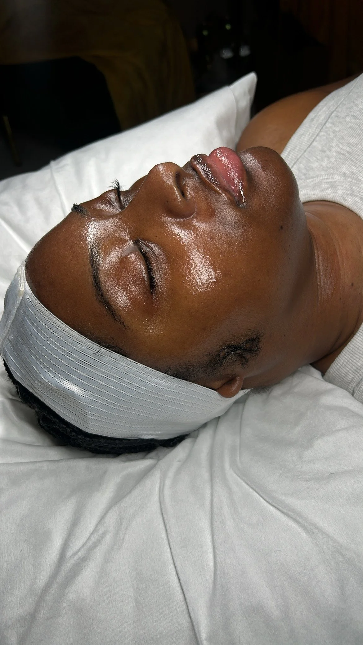 A person lying down with their eyes closed, wearing a white head wrap, gold chain, and a gray shirt, with a close-up of their face showing smooth skin and a nose piercing.