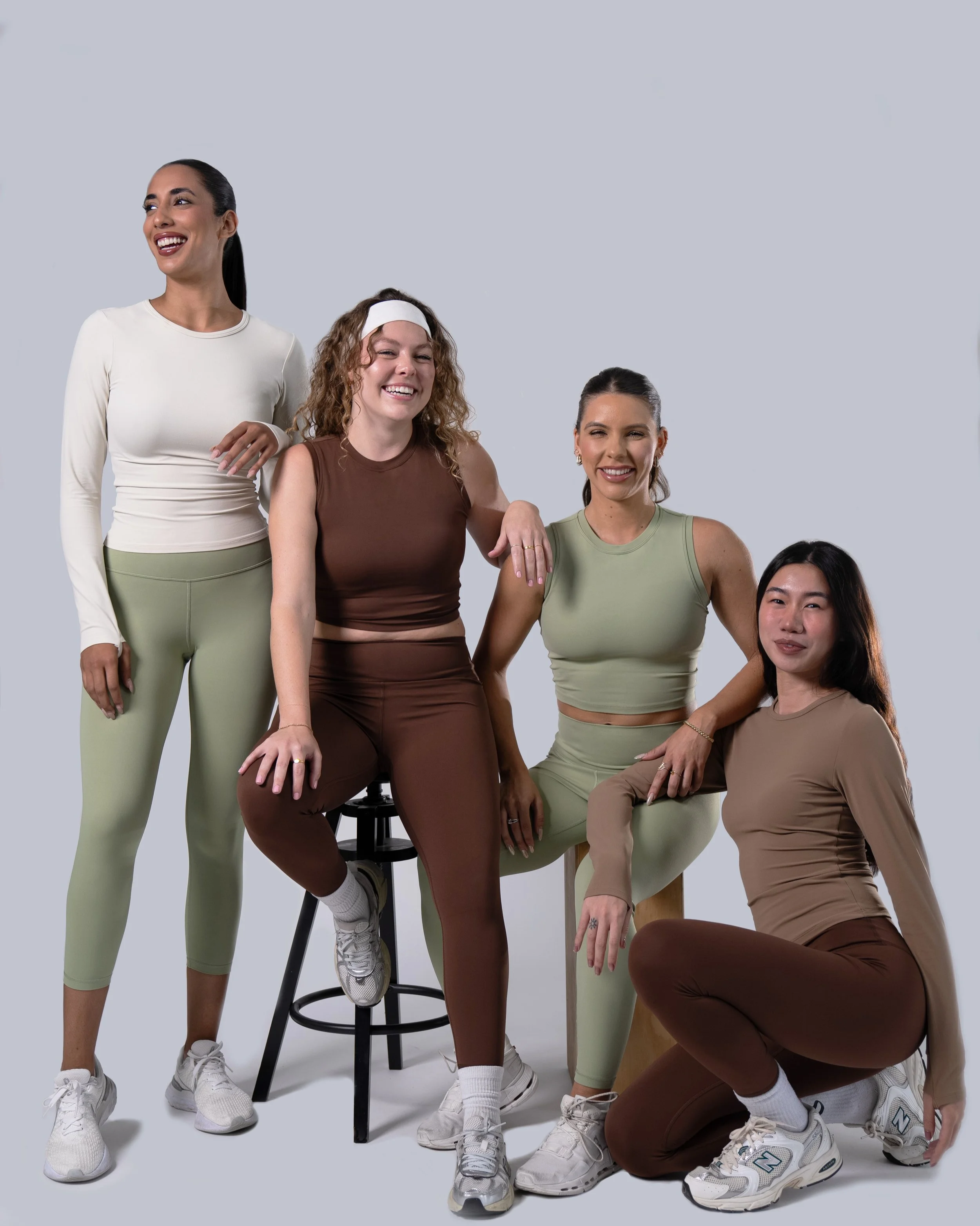Four women dressed in athletic wear smiling and posing together in a studio with a plain background.