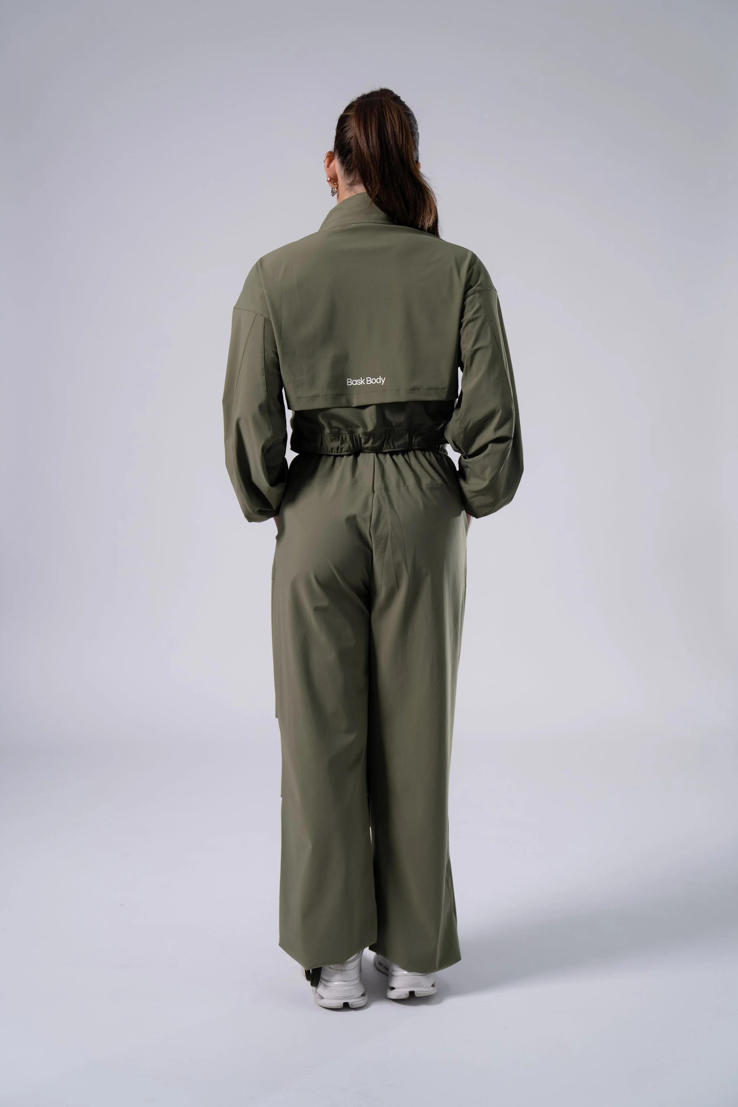 Back view of a woman in a green tracksuit with a cropped jacket and wide-leg pants, standing against a plain light gray background, wearing white sneakers.