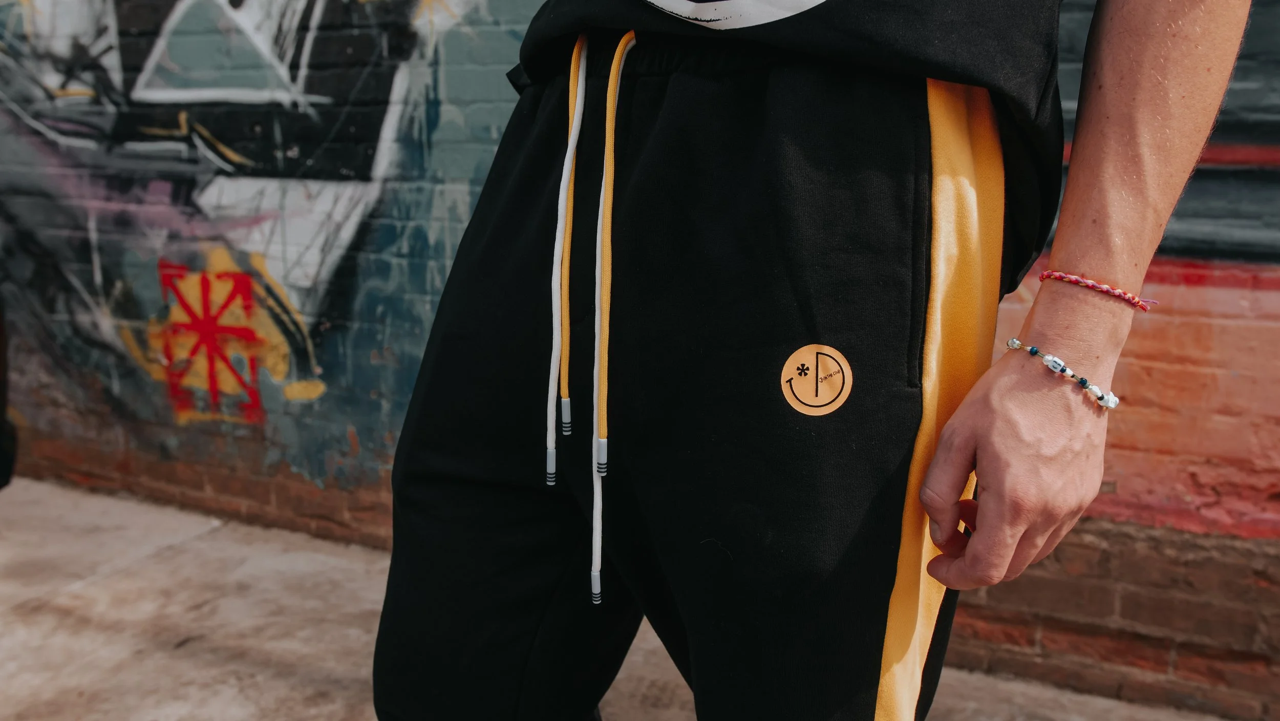 Person standing outdoors wearing black and yellow pants with a branded logo on the pocket, a dark T-shirt, and a beaded bracelet on their wrist, against a brick wall with graffiti.