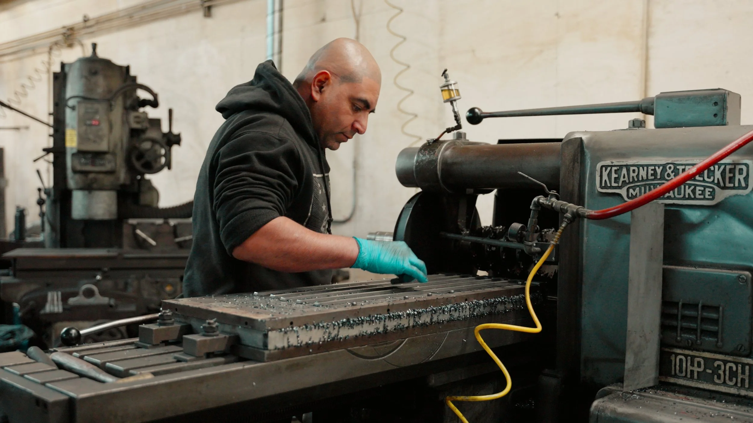 Man wearing a black hoodie and blue gloves operating a milling machine in an industrial workshop.