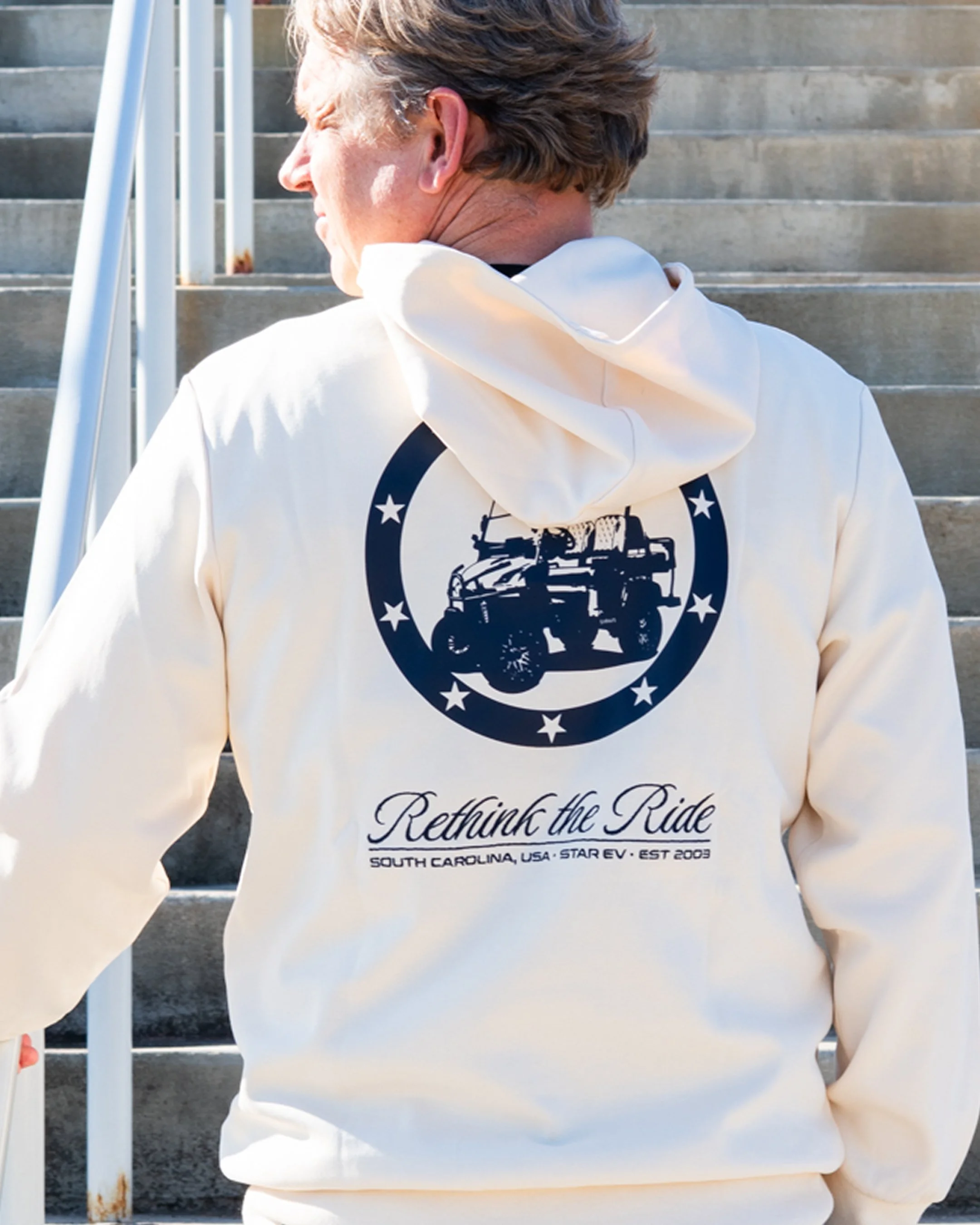 A person with short brown hair wearing a cream hoodie with a logo of a military tank and stars, standing on stairs outside, with text that reads 'Rethink the Ride' and 'South Carolina, USA, Star EV, Est 2003.'