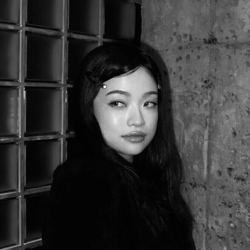 A woman with dark hair styled with hair clips, looking to the side, standing against a textured concrete wall and a glass block wall, in black and white.