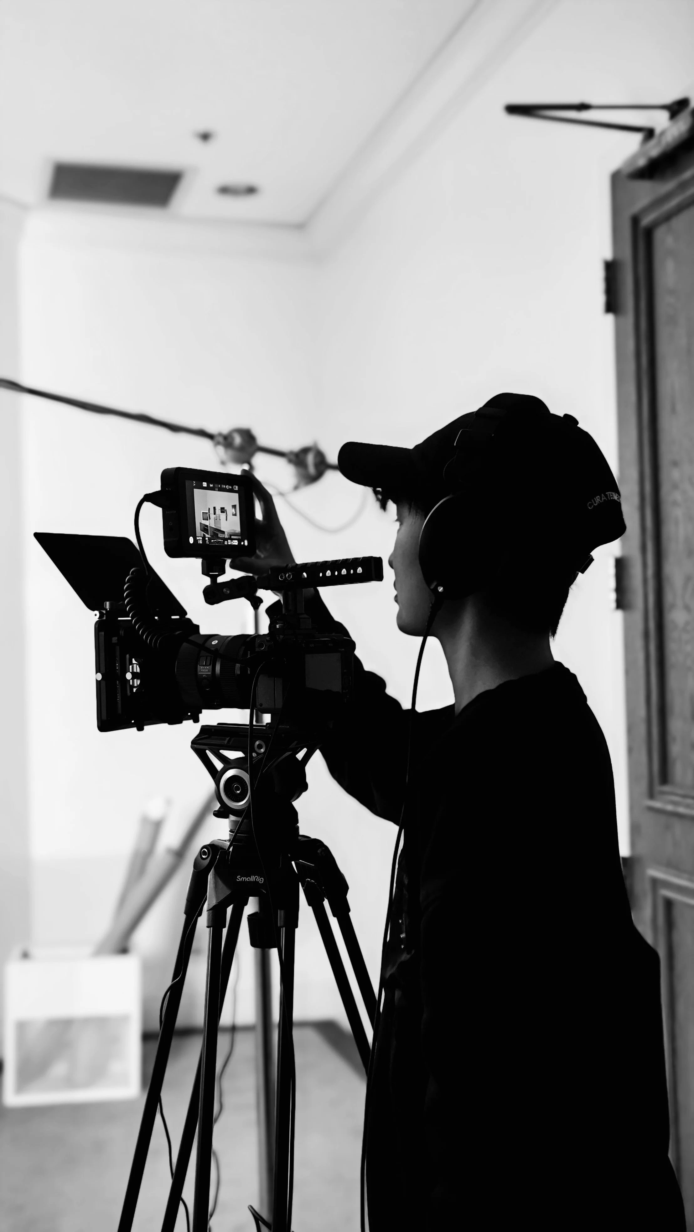 Person operating a video camera on a tripod, wearing headphones, in an indoor setting.