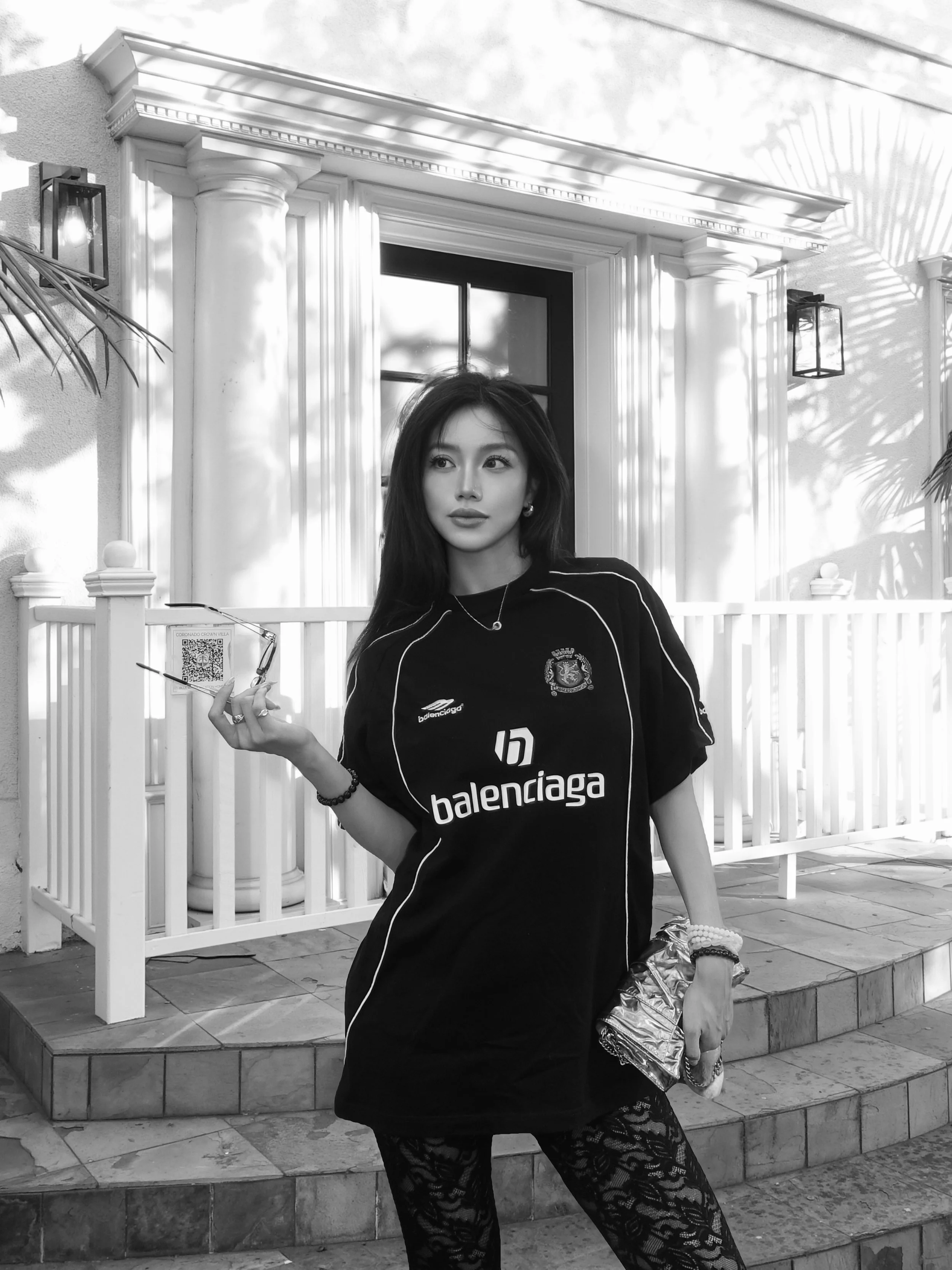 A woman with long dark hair standing outdoors in front of a white picket fence and a house with a window and columns. She is wearing a black sports jersey with logos, patterned lace pants, and jewelry, holding a pair of eyeglasses and a shiny clutch bag.
