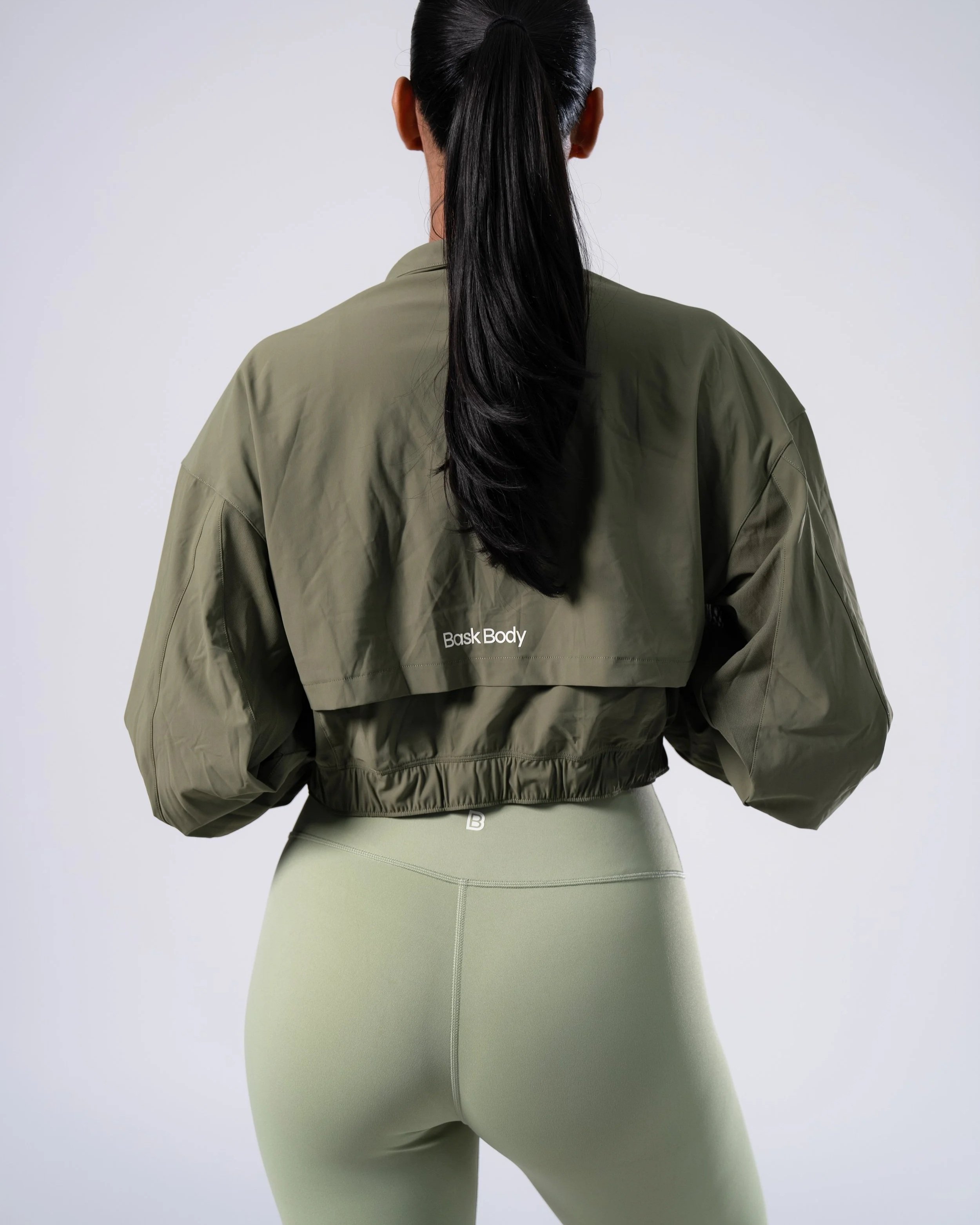 A woman is standing with her back facing the camera, wearing a Bask Body UPF 50 green jacket and light green leggings, with her long black hair tied in a ponytail, against a plain white background.