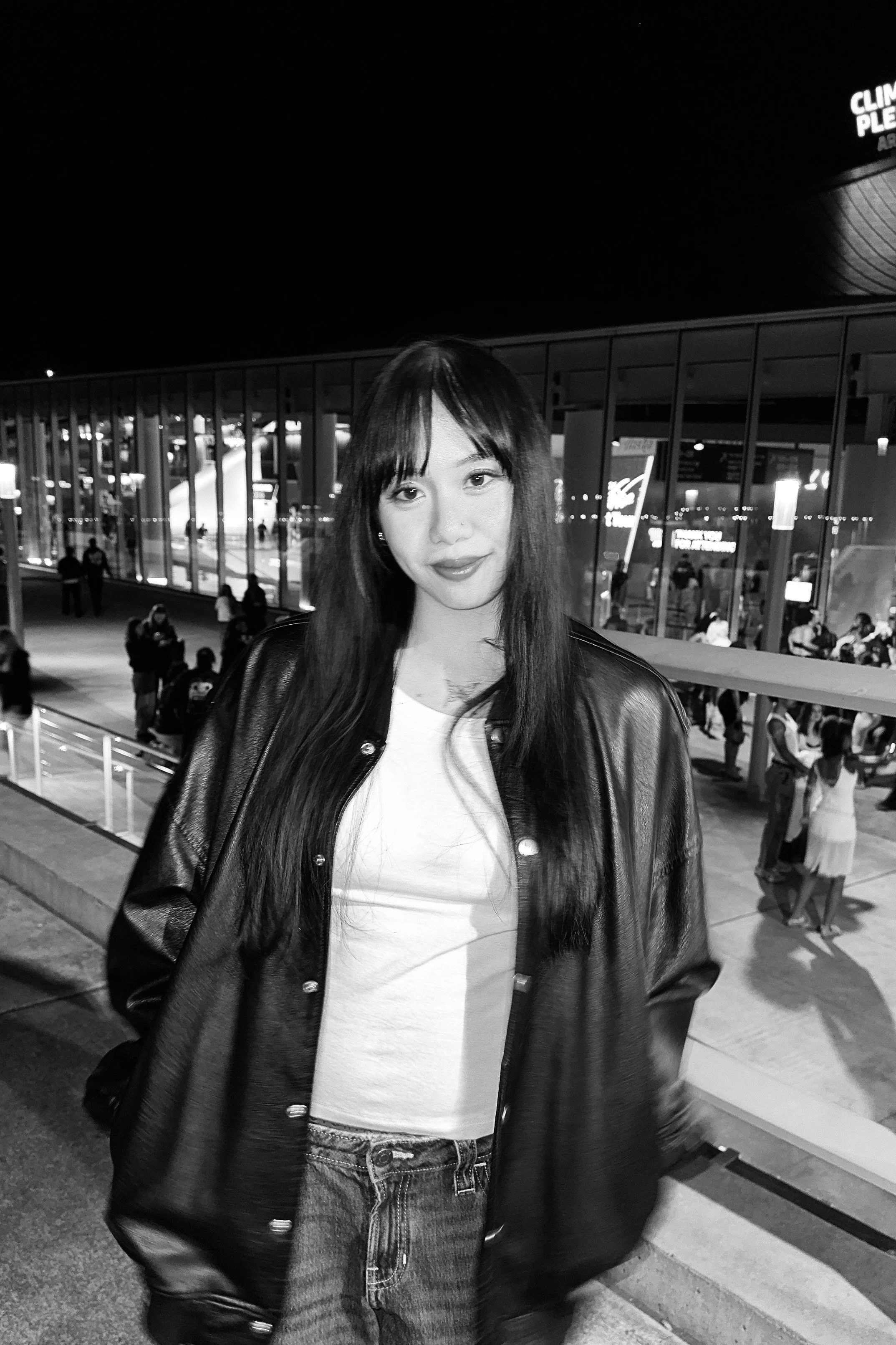 A young woman with long dark hair and bangs standing outdoors at night, wearing a leather jacket and white top, near a glass building with people in the background.