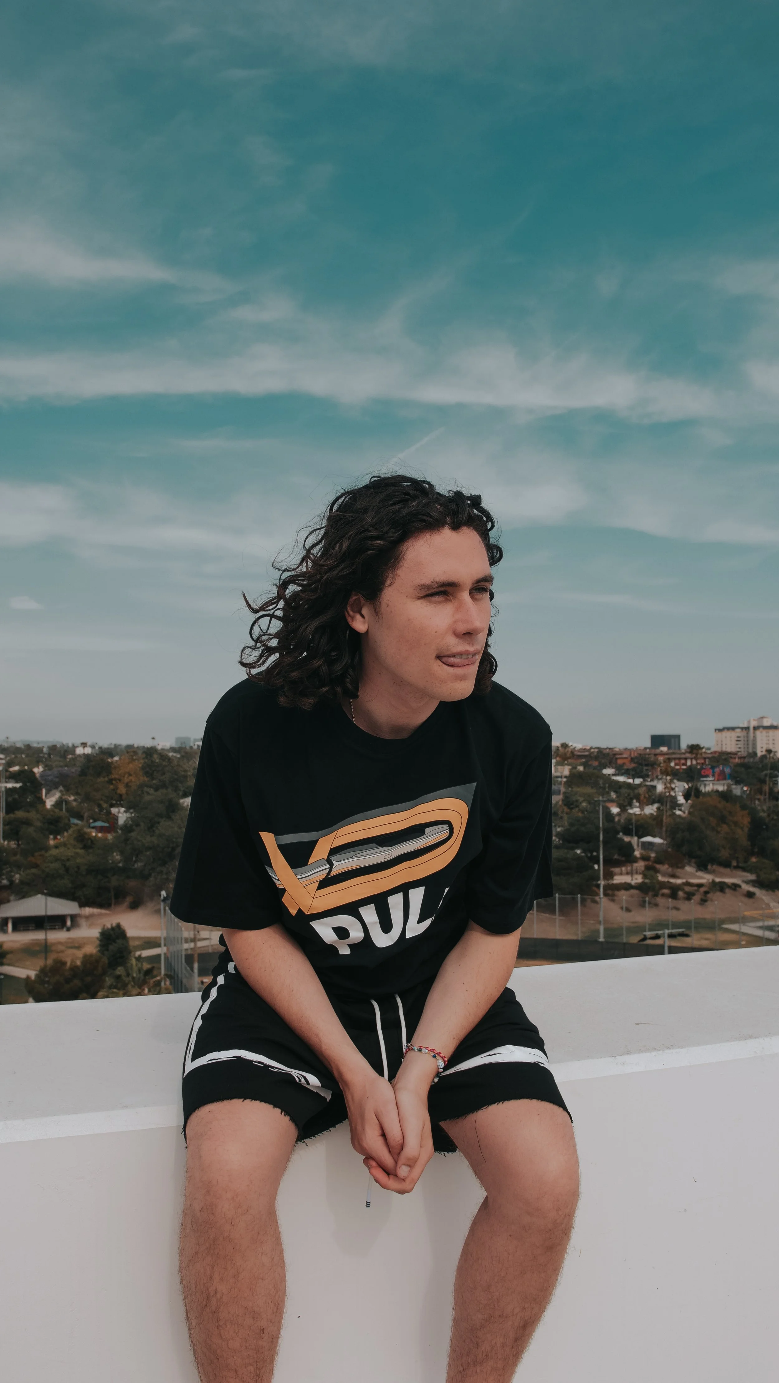 Young man with long curly hair sitting on a white ledge outdoors, wearing a black t-shirt with a logo and shorts, with a cityscape and blue sky with clouds in the background.