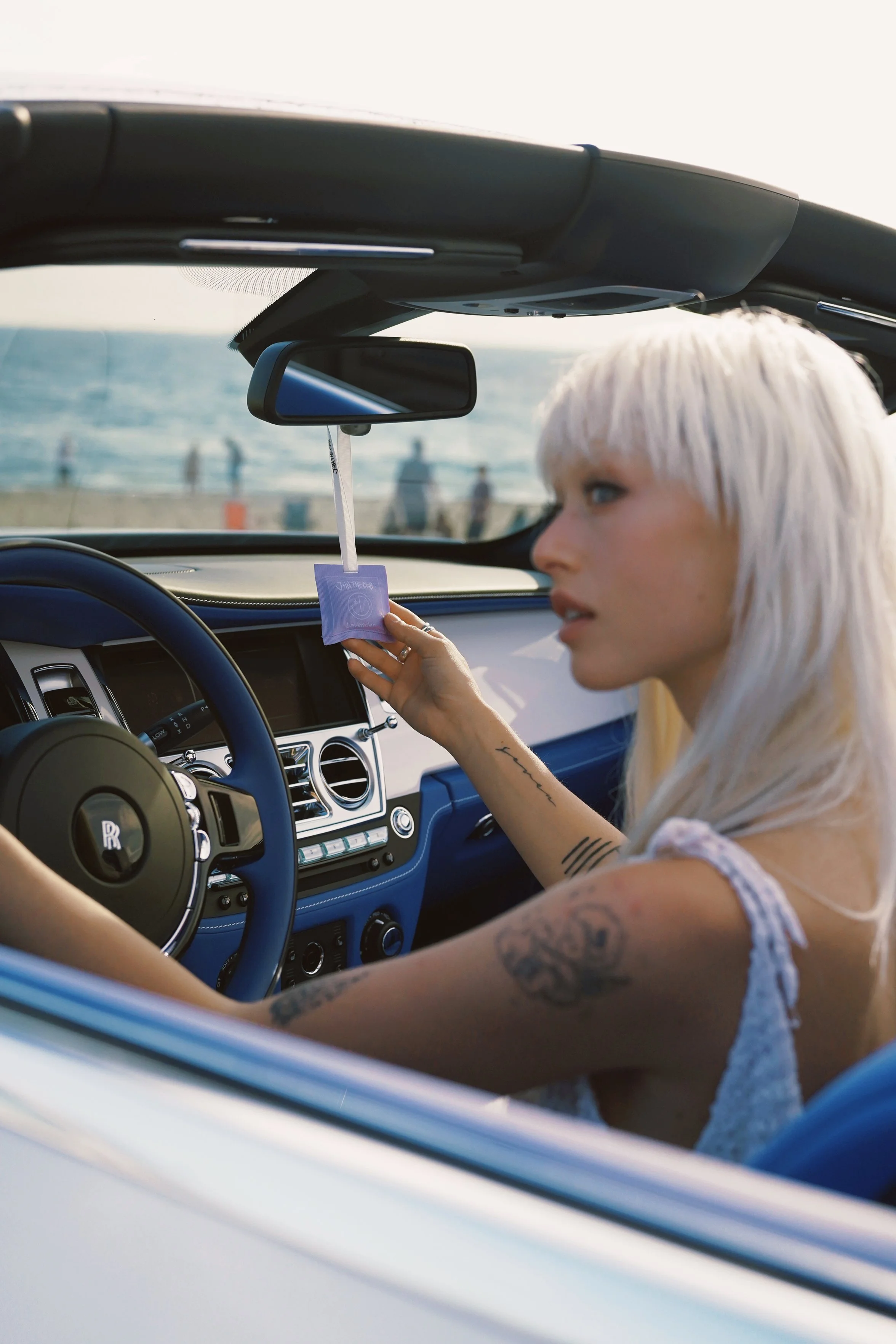 A woman with platinum blonde hair and tattoos on her arm sitting in the driver's seat of a white convertible car near the beach, holding a lavender air freshener.