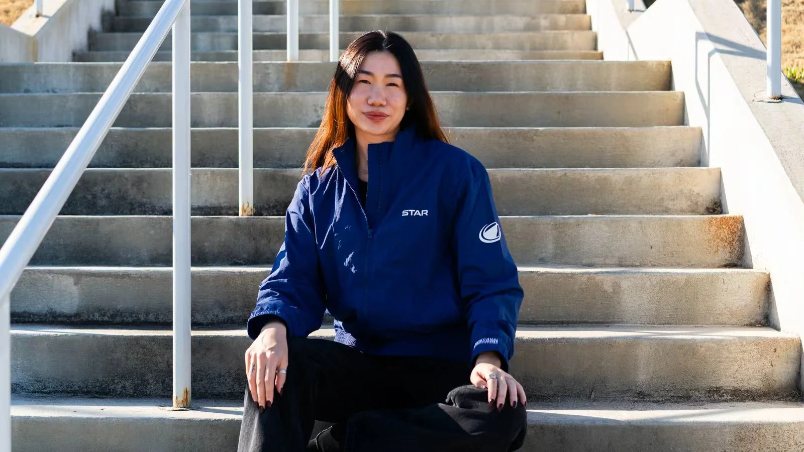 A woman sitting on outdoor stairs with metal railings, wearing a blue jacket with logos, smiling, with sunlight highlighting her face.