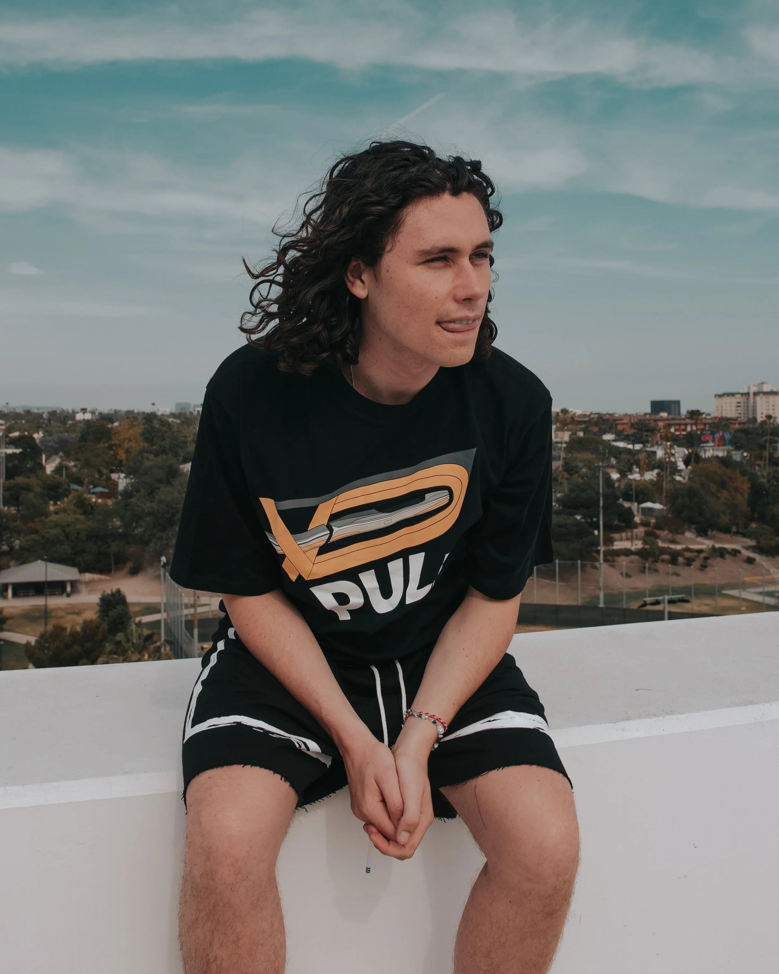 Young man with curly hair and freckles, wearing a black t-shirt with a Superman logo, sitting outdoors against a cityscape background with cloudy sky, showing his tongue and hands clasped.