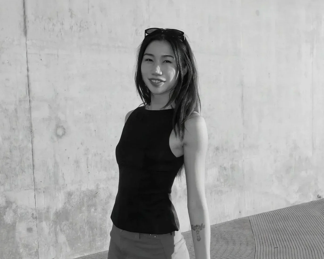 A young woman with long dark hair, wearing a black sleeveless top and pants, posing with a smile in front of a textured concrete wall, with sunglasses on her head and a floral tattoo on her left arm.