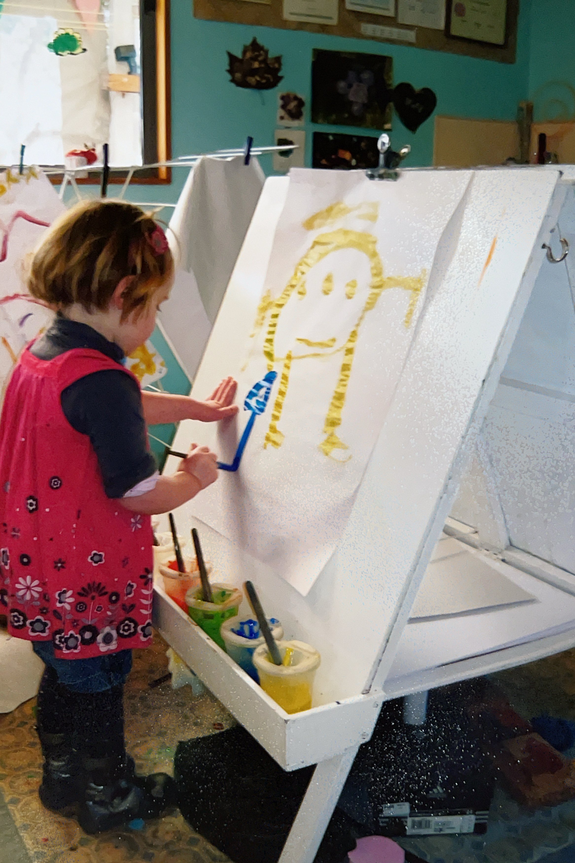 A young girl painting a portrait on an easel with colorful paints and brushes, in an art classroom.