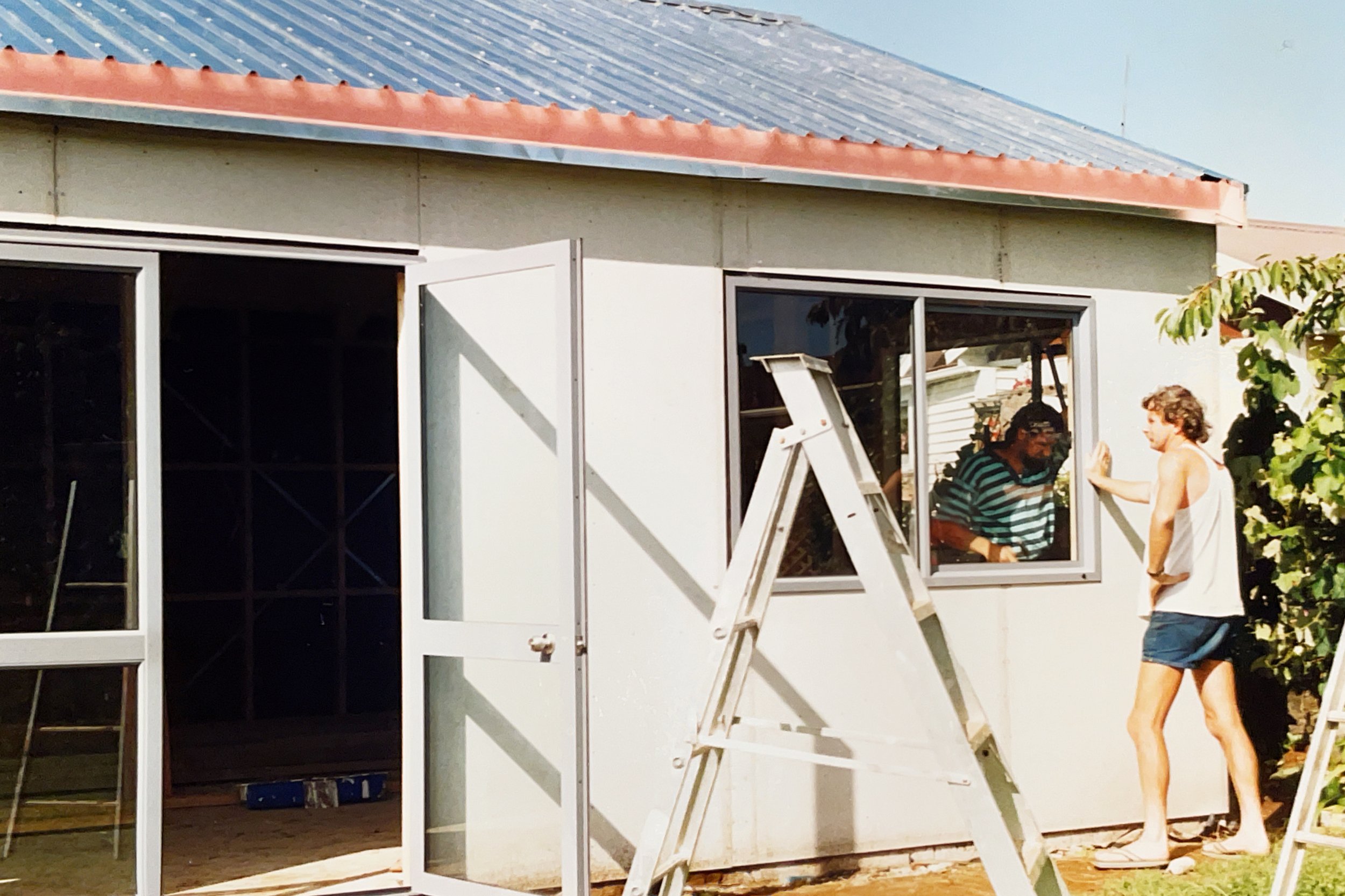 Two women building a house, one inside and one outside, with a ladder and construction materials visible.