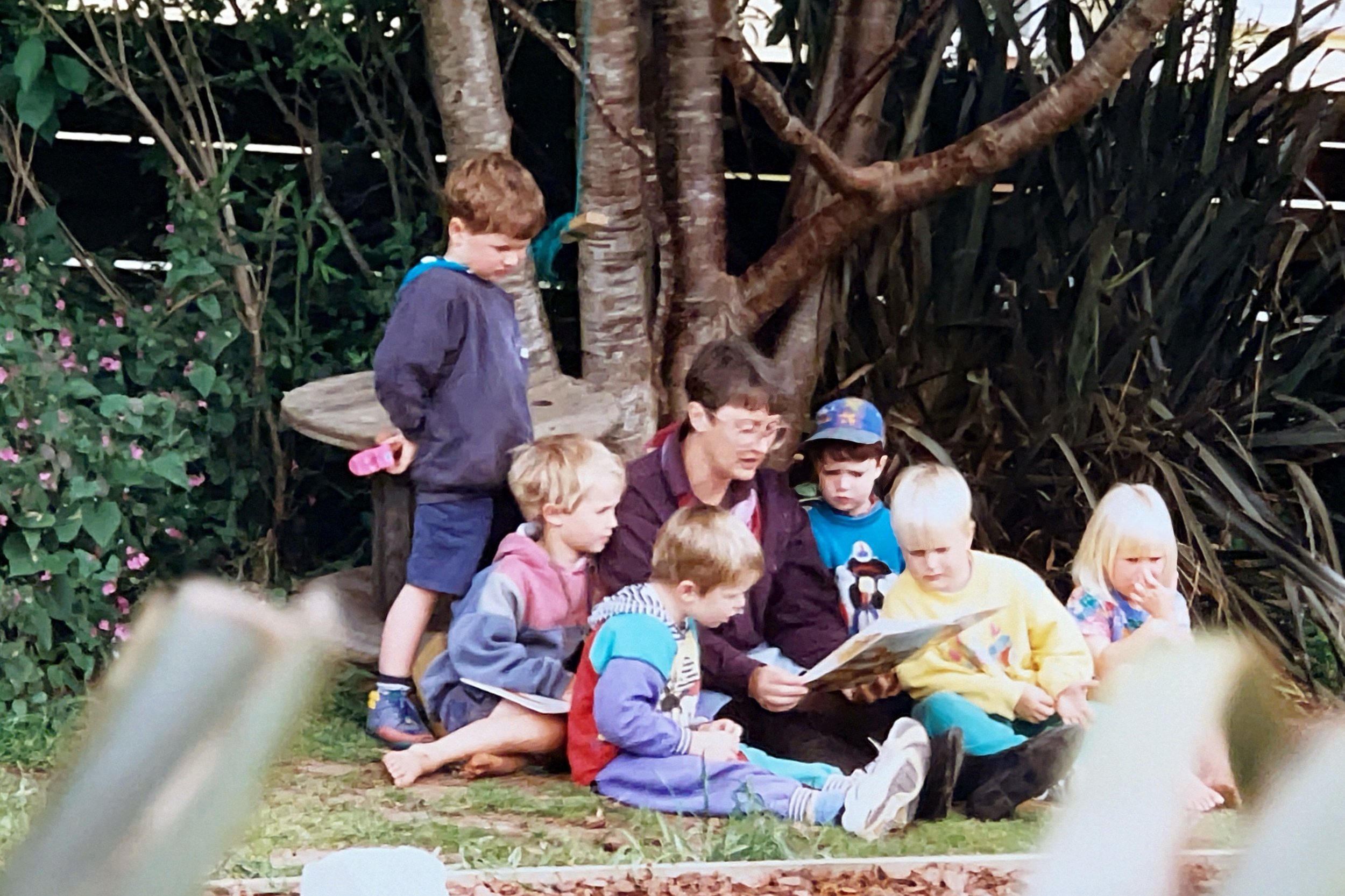 A woman with glasses reading a book to seven children sitting and standing outdoors near a tree, surrounded by green foliage.