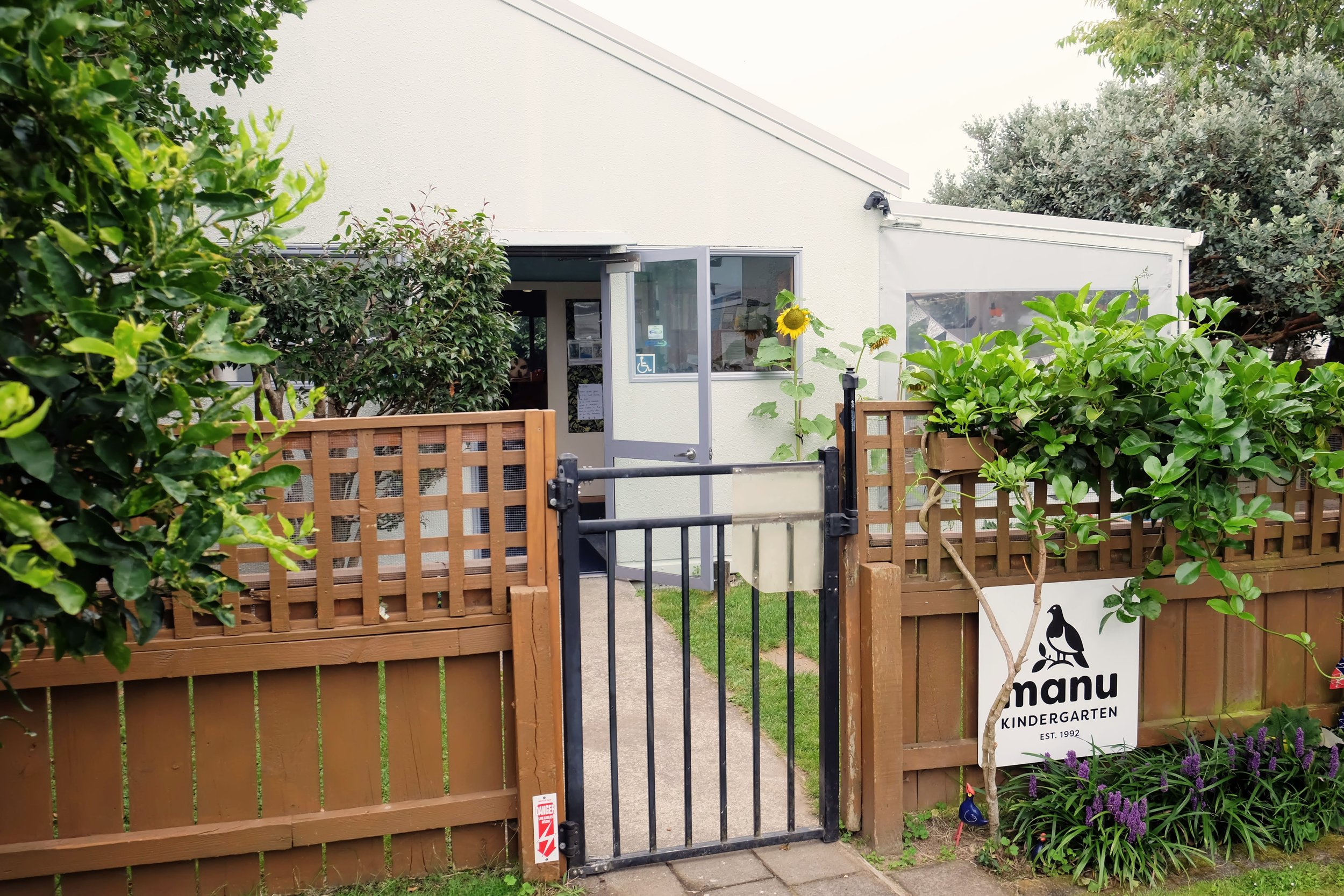 A single-story building surrounded by a wooden fence and gate, with a sign labeled "manu Kindergarten EST. 1992." The area is landscaped with grass, trees, and shrubs.