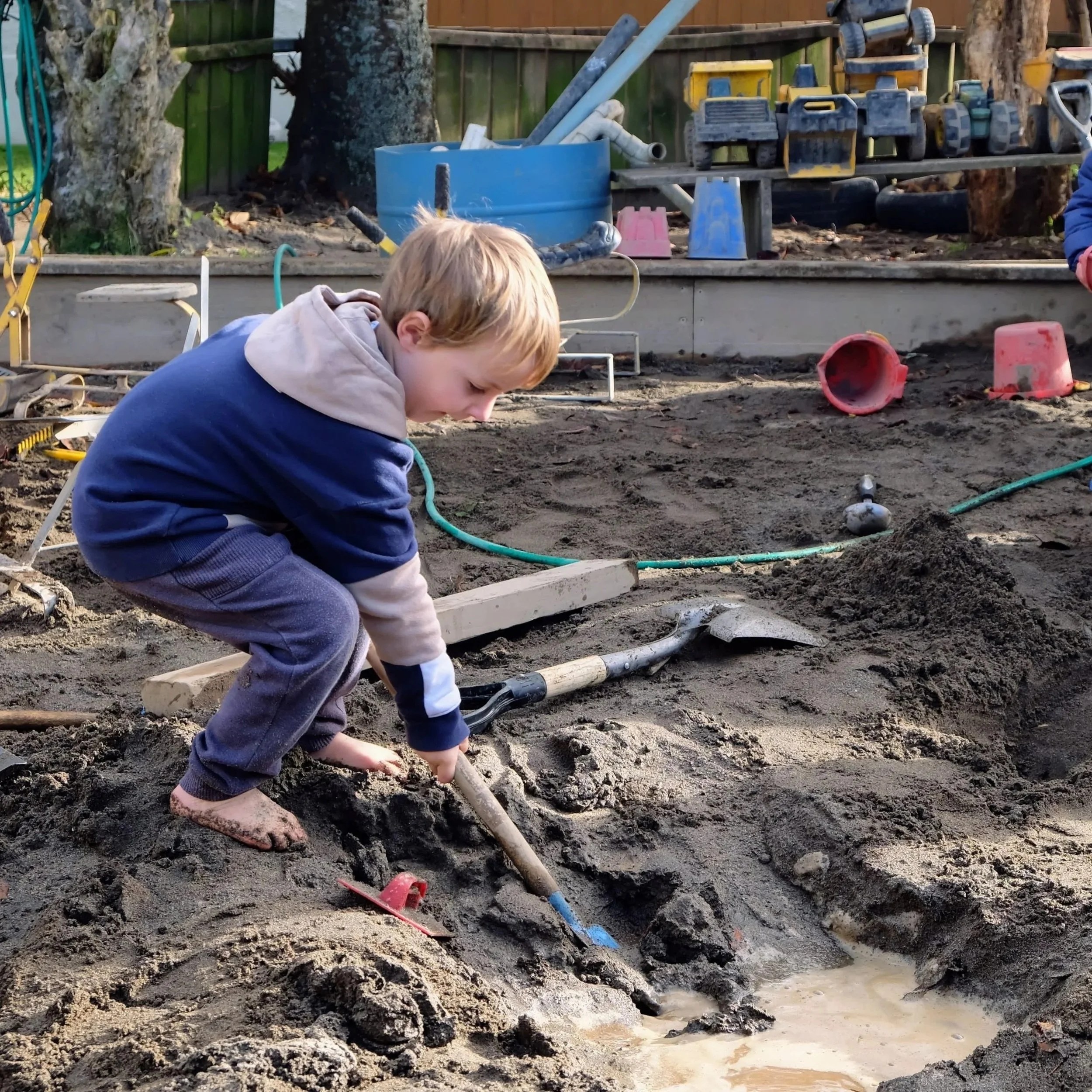 A young boy with blond hair, wearing a blue and gray hoodie and gray pants, is playing in a sandbox with a small shovel, digging in the dirt.