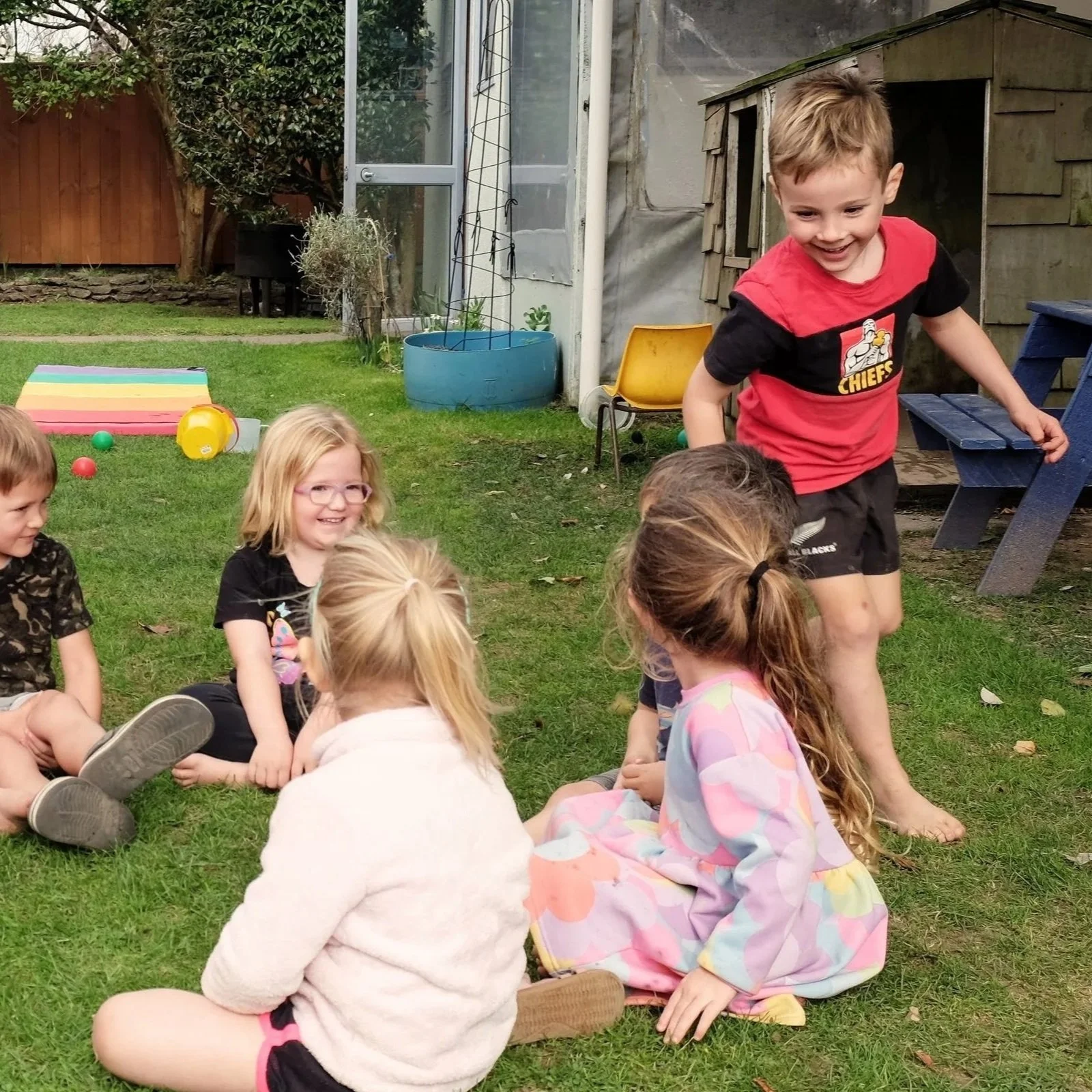 Children playing outdoors on the grass, sitting in a circle, with one child smiling and standing barefoot nearby.