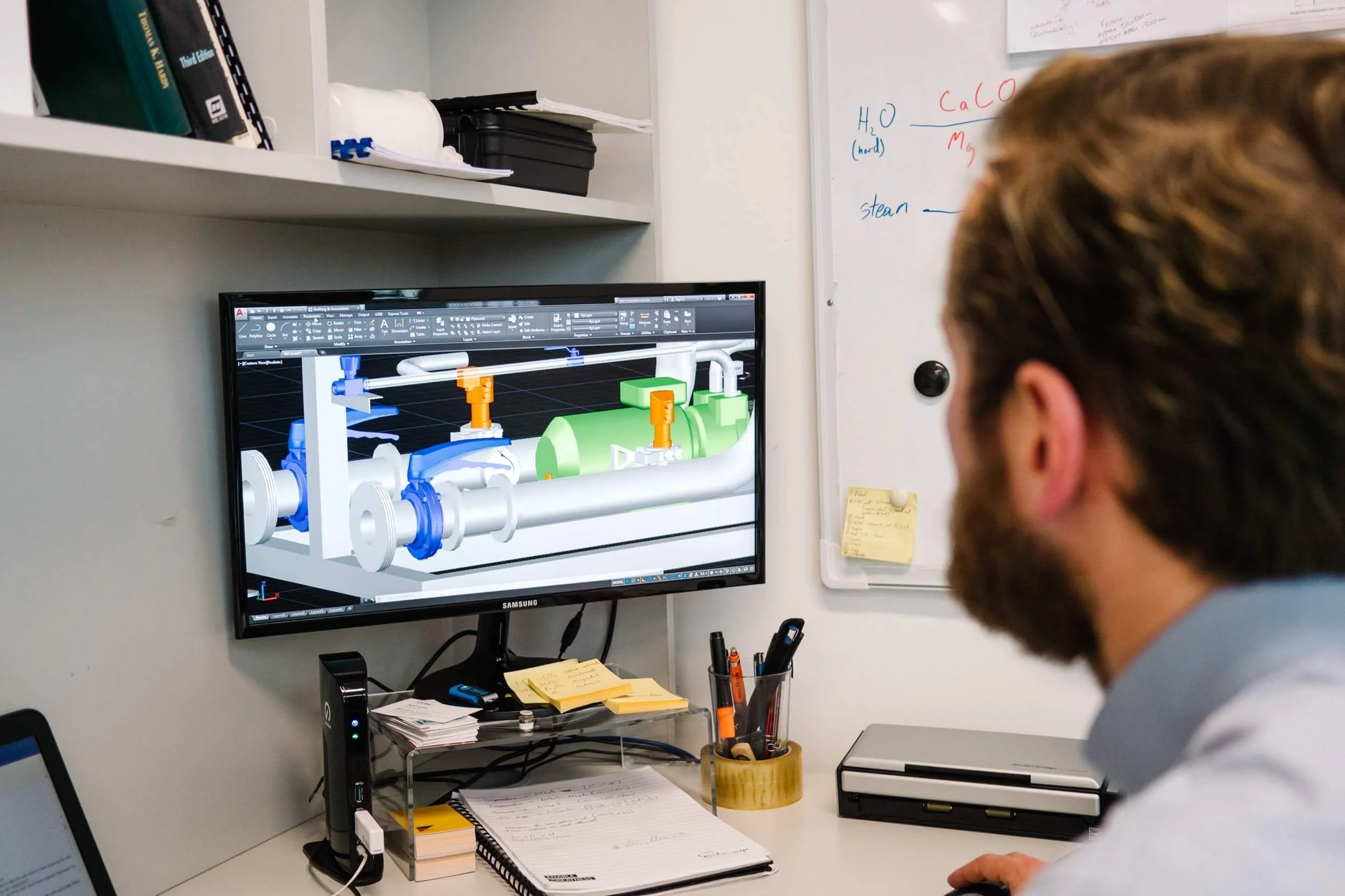 A person watching a computer monitor displaying a 3D design of mechanical piping and valve systems in an office or workspace with a whiteboard and desk accessories.