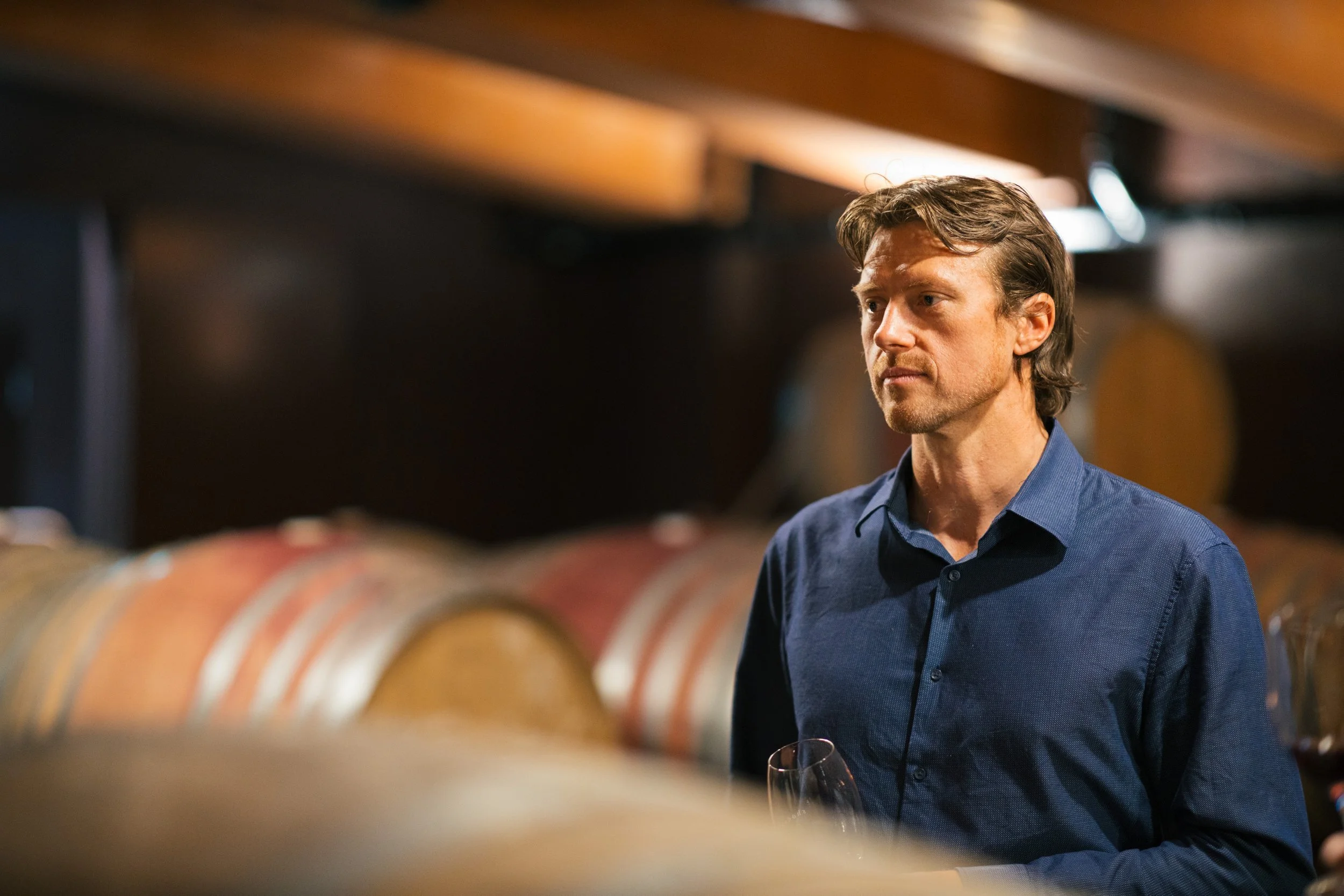 Man in a blue shirt standing in a wine cellar with wooden barrels.