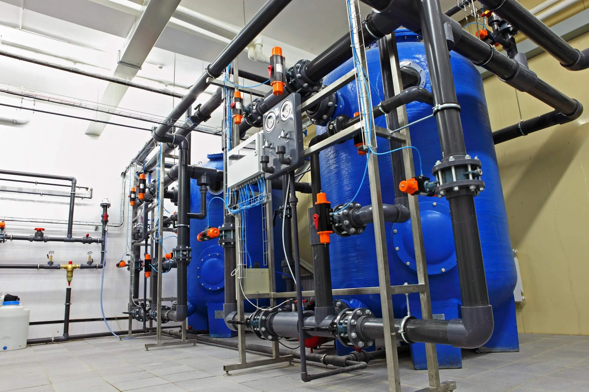 Industrial water treatment equipment with large blue tanks, black pipes, and orange valve handles in a clean, well-lit room.