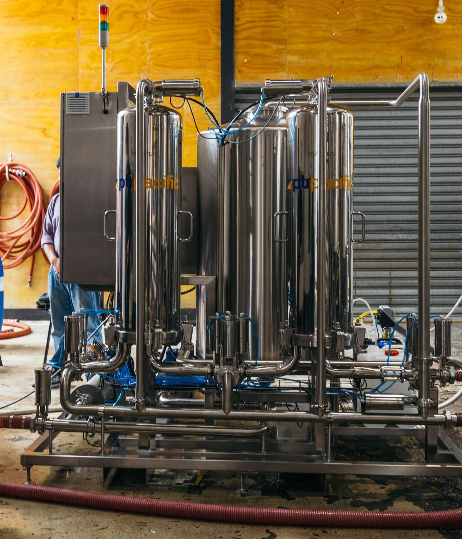 Industrial brewing equipment in a brewery with stainless steel tanks and pipes, a control panel, and a worker partially visible in the background.