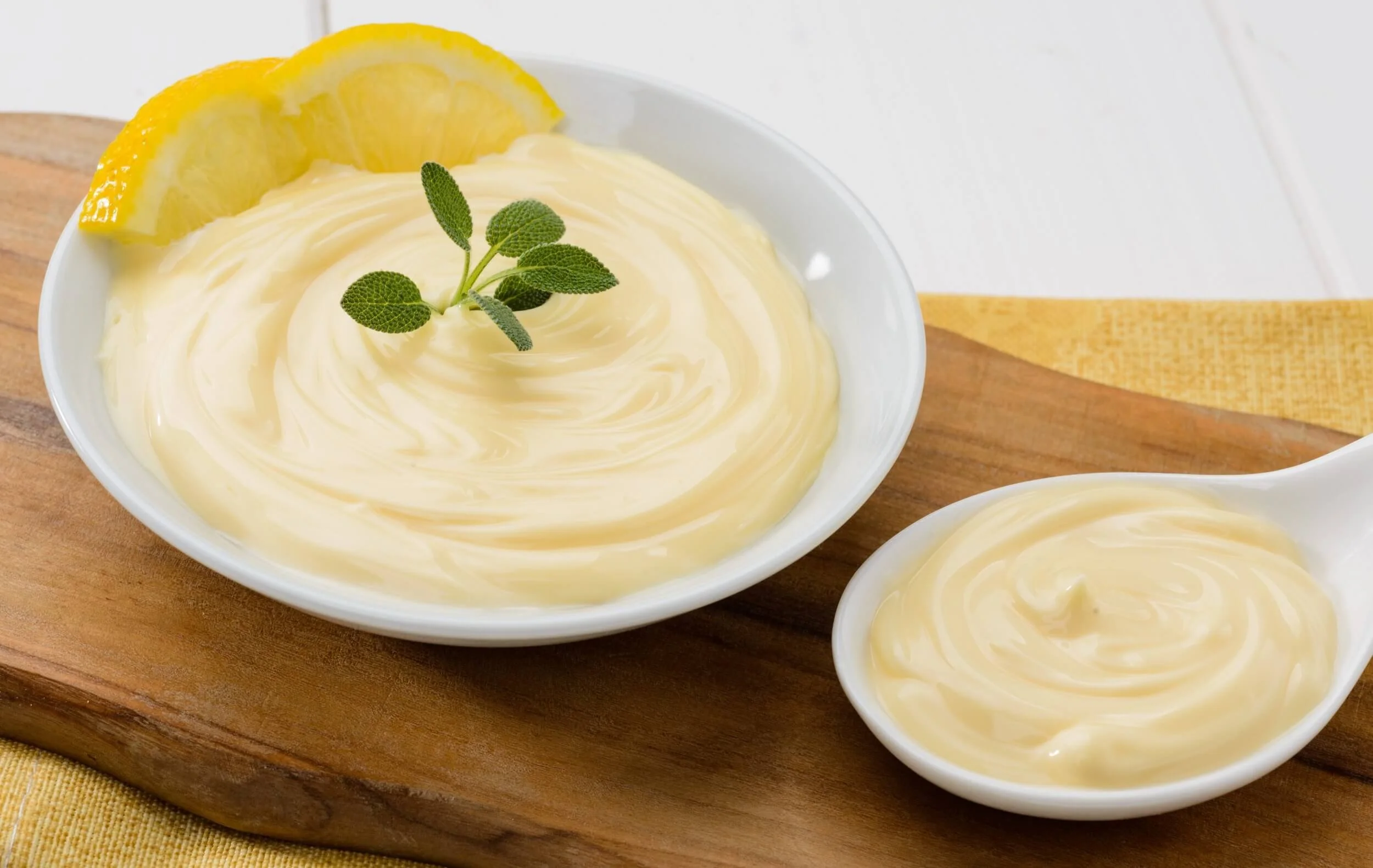 A bowl of lemon-flavored creamy dessert garnished with lemon slices and a sprig of mint, placed on a wooden serving board with a side of more dessert in a small spoon.