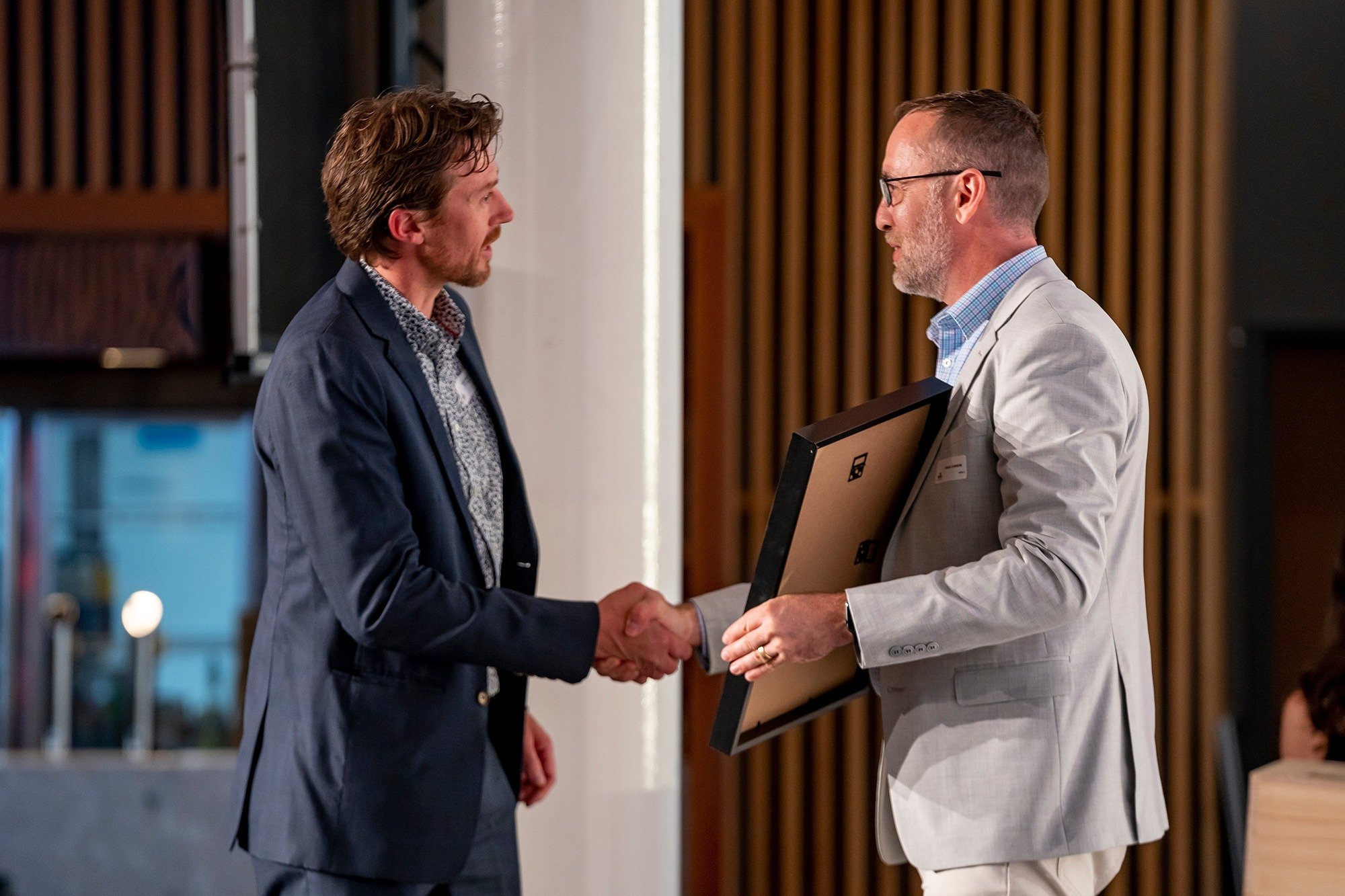 Two men shaking hands, one holding a framed certificate in a professional setting.