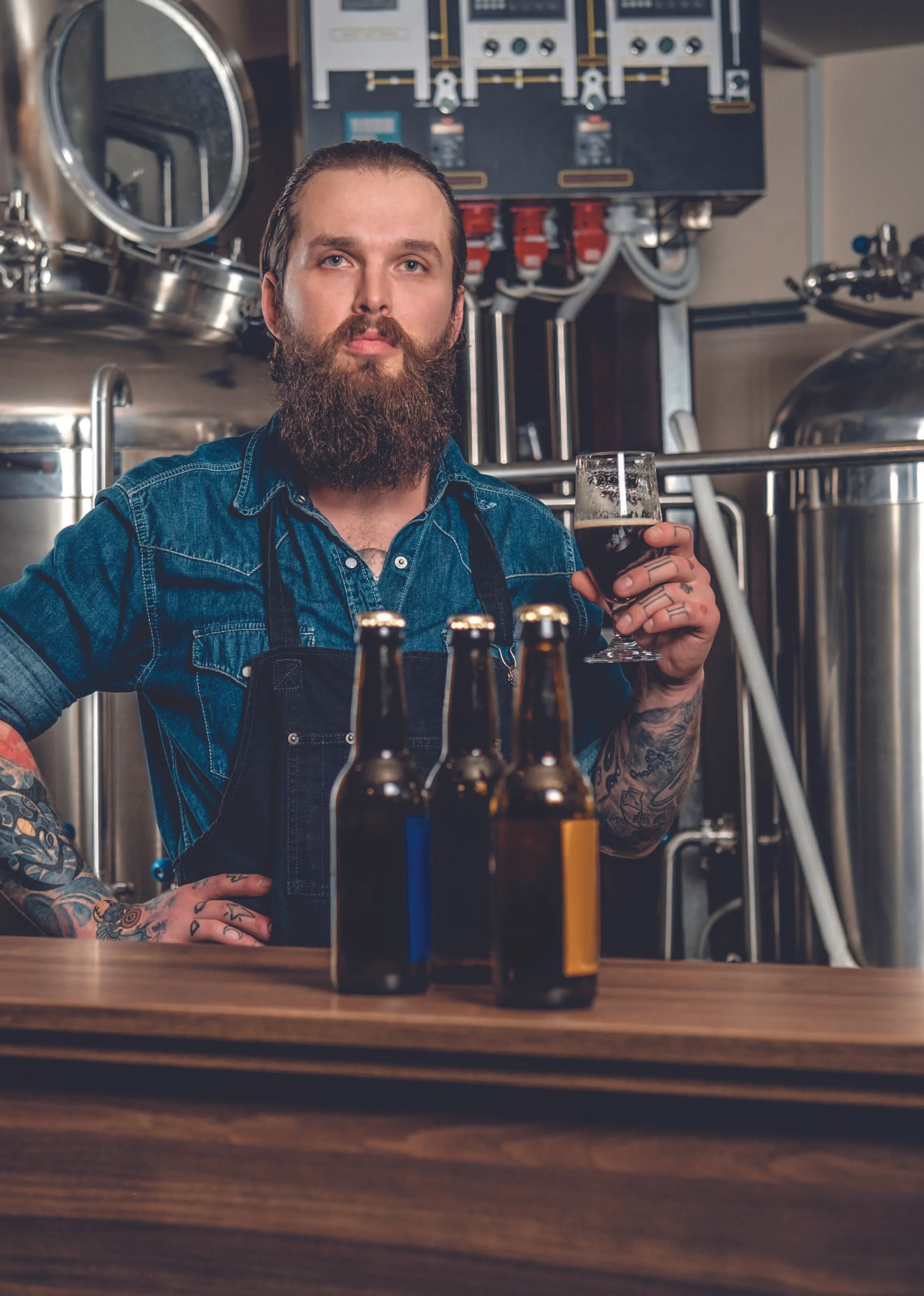 A bearded man with tattoos, wearing a denim shirt and black apron, holding a glass of dark beer in a brewery with large stainless steel brewing tanks and equipment in the background.