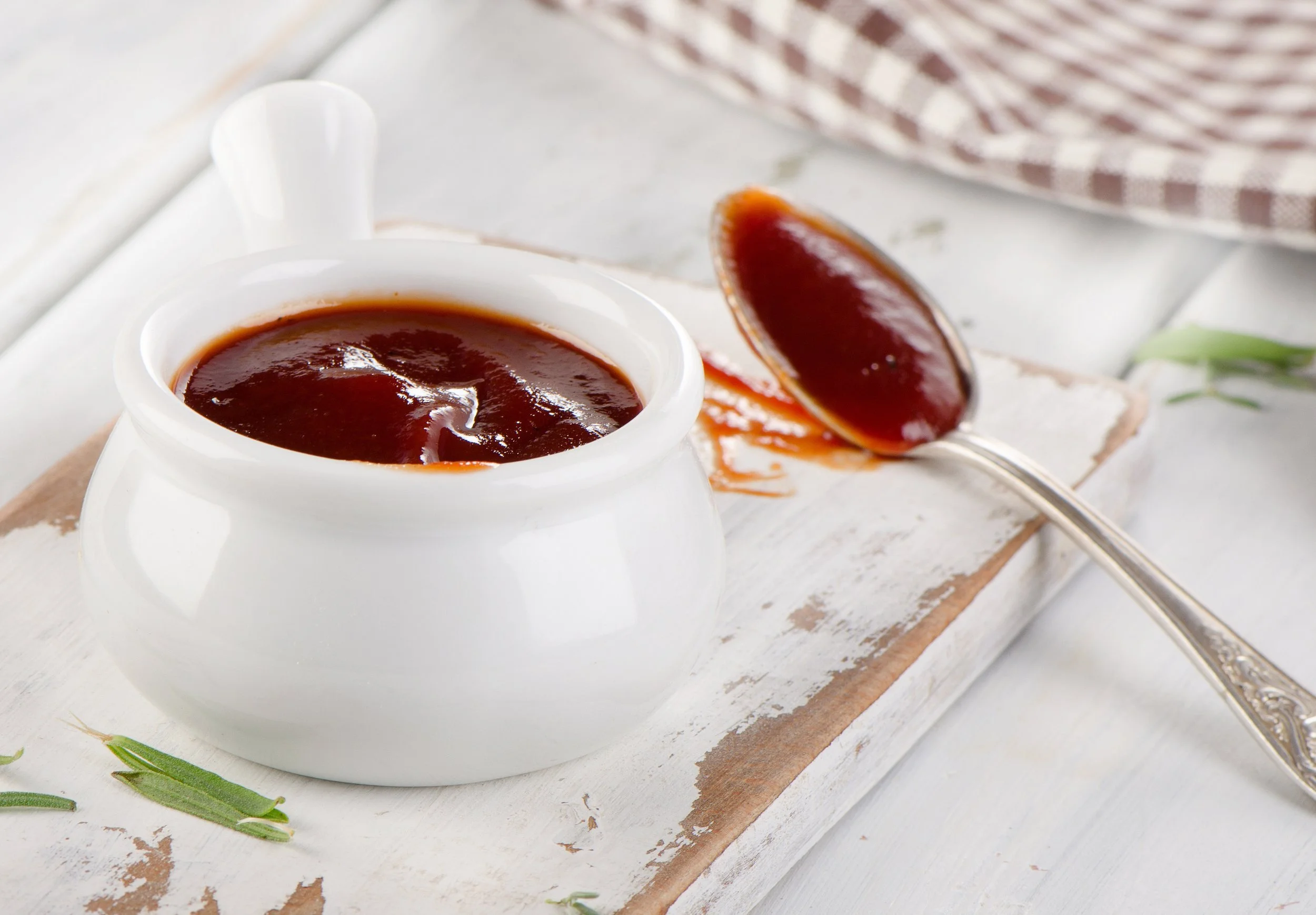 A white ceramic pot filled with barbecue sauce, with a silver spoon coating with sauce resting next to it on a rustic white wooden board, with some rosemary sprigs nearby.