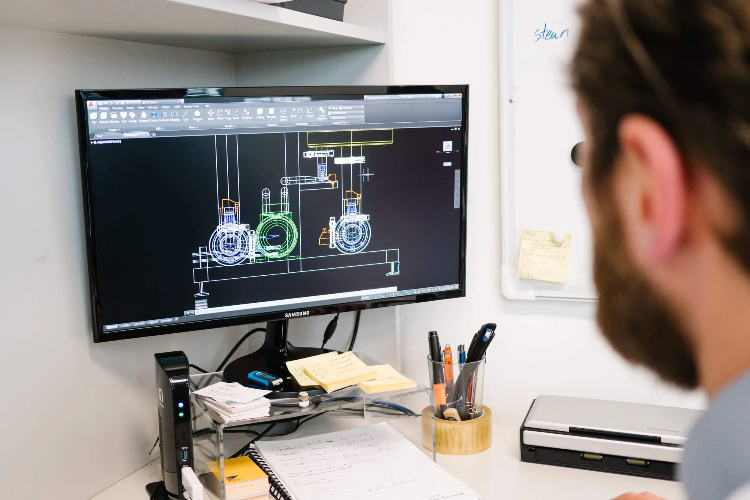 Person working on CAD design on a computer monitor, surrounded by pens, notes, and a whiteboard with writing.