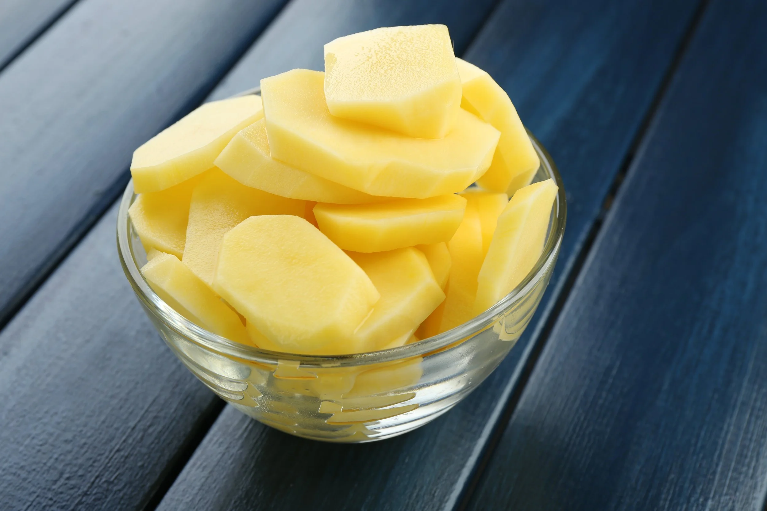 A glass bowl filled with peeled, sliced yellow potatoes on a dark blue wooden surface.