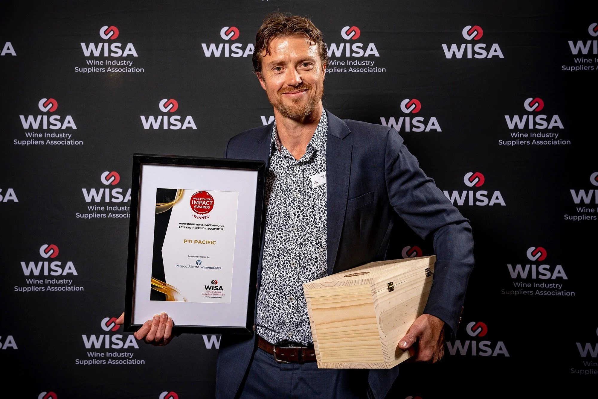 Person holding an award plaque and wooden box, standing in front of a "Wine Industry Suppliers Association" backdrop at an event.